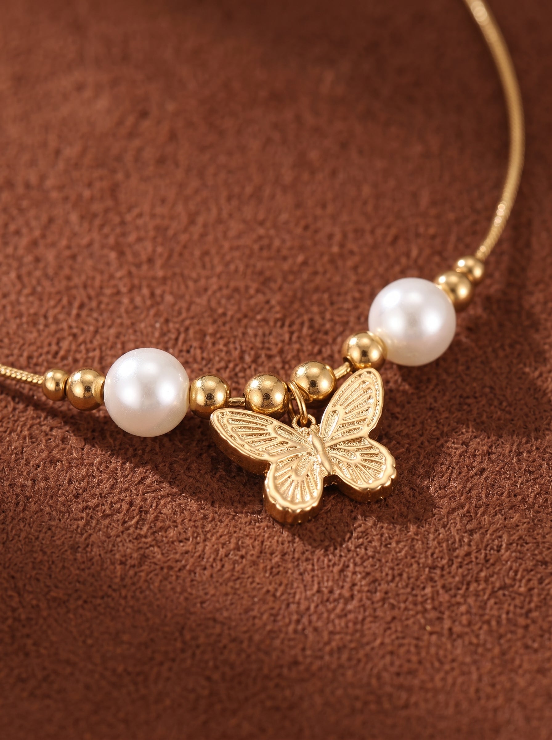 Gros plan du collier Ephemeral Dance mettant en valeur un pendentif papillon doré avec deux perles blanches d'eau douce et des perles dorées sur une fine chaîne dorée, présenté sur une surface texturée marron. @pearl