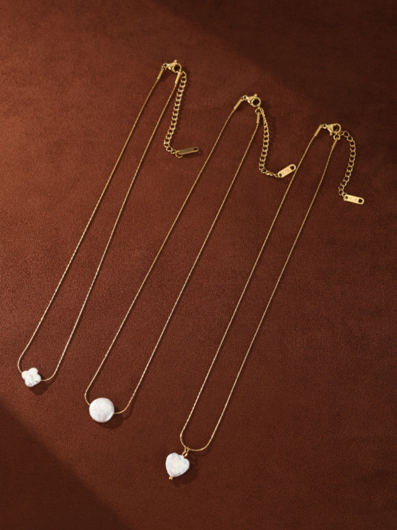 Trois colliers ton or, chacun avec un unique pendentif en perle d'eau douce : un quatrefeuille, une perle ronde, et une perle en forme de cœur, présentés sur une surface texturée marron.