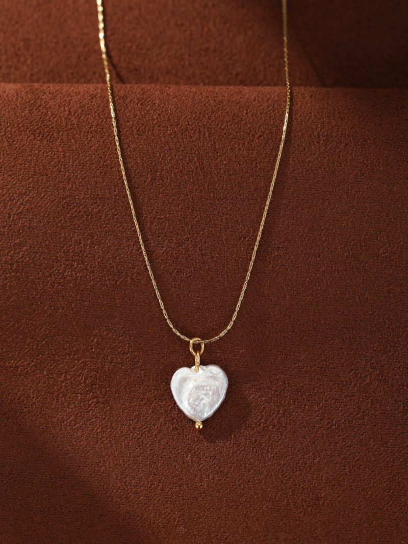 Un délicat collier à chaîne ton or présentant une seule et lustrée perle blanche d'eau douce en forme de cœur, accentuée d'une minuscule perle dorée en bas. Le collier est présenté sur fond marron riche et texturé.