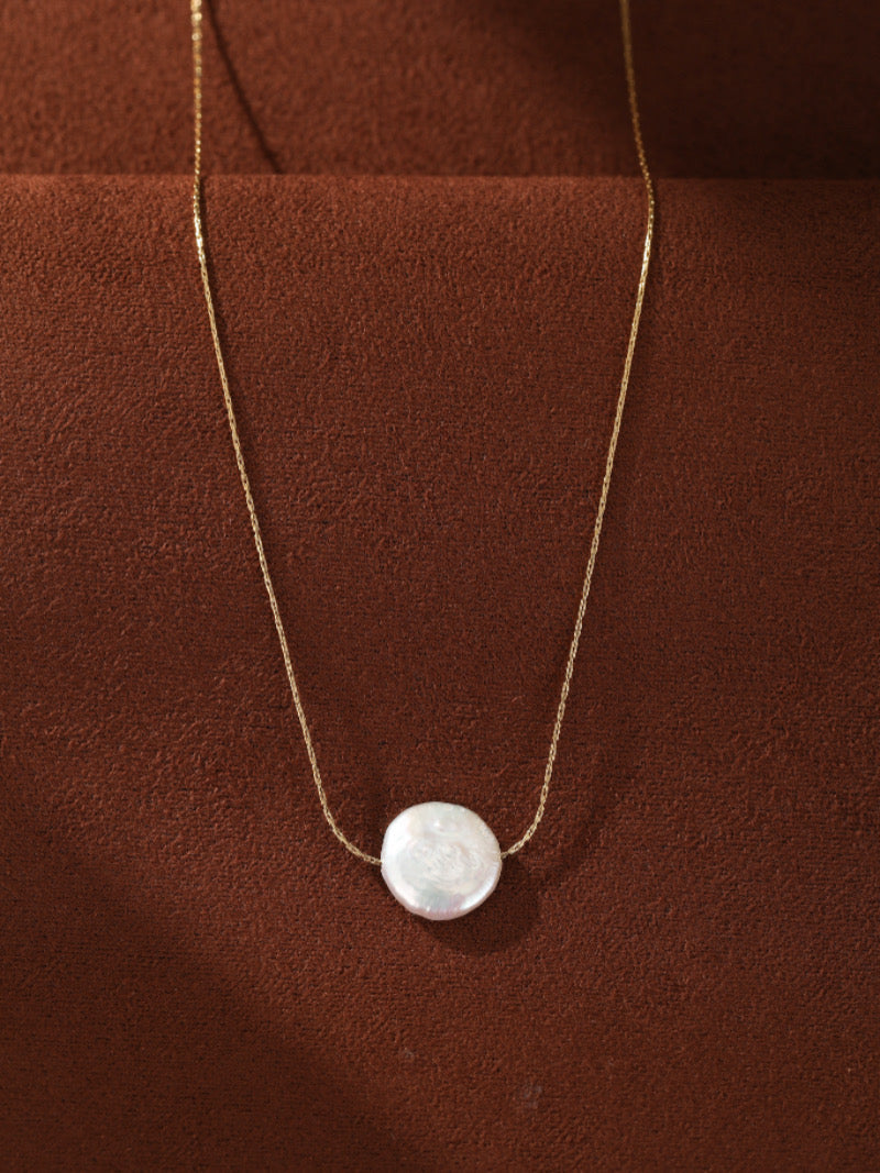 Collier pendentif cœur quatrefeuille Essentia Form en perle d'eau douce présentant une seule perle blanche plate et ronde sur une délicate chaîne dorée contre un fond texturé marron.