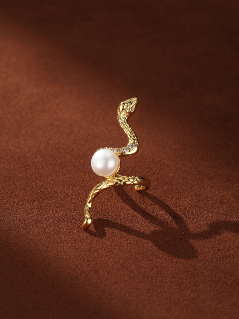 Gold snake cuff ring featuring a white freshwater pearl and subtle accent stones on the snake's body, set against a textured brown background. @pearl