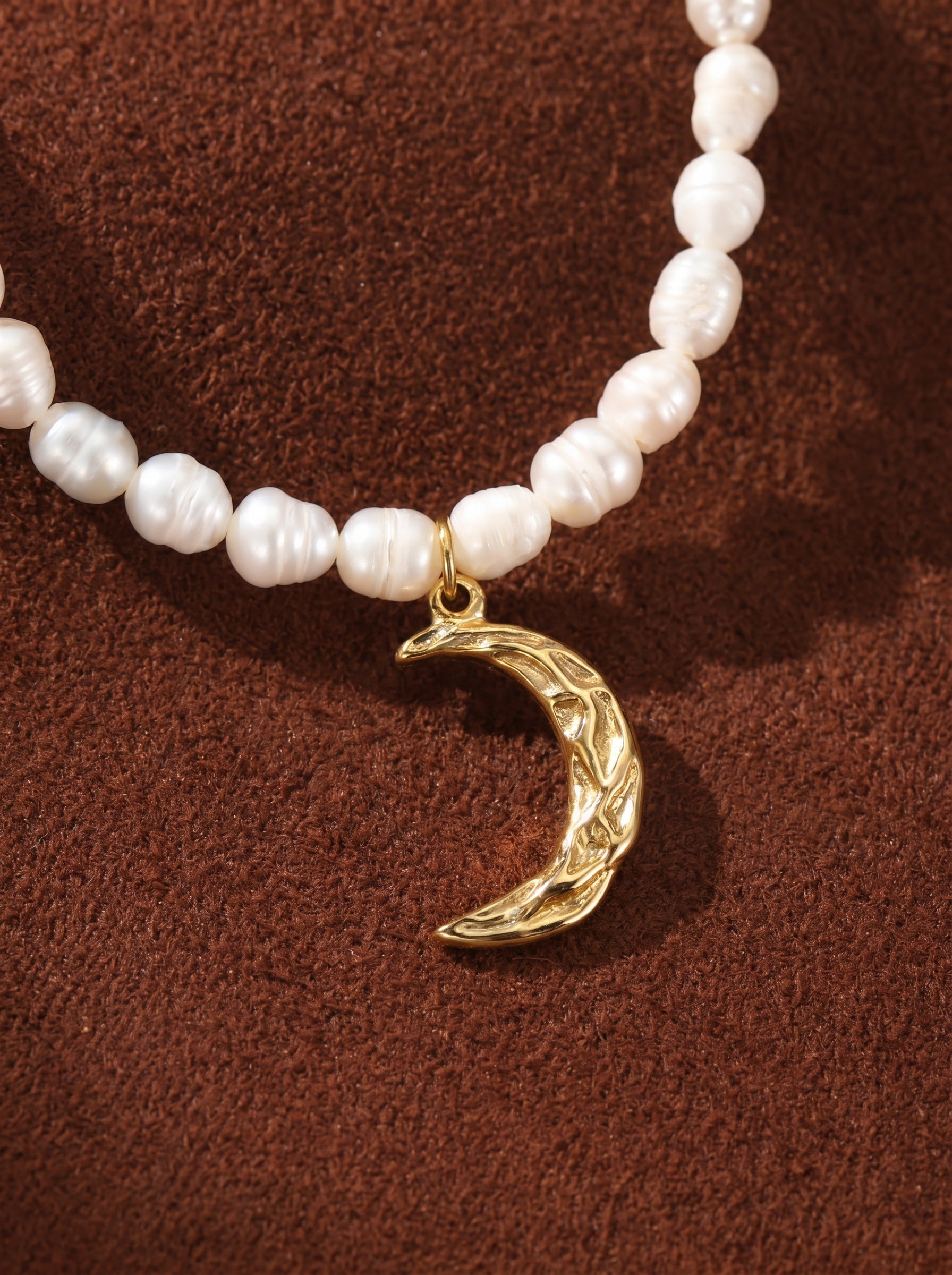 Close-up of a delicate choker necklace featuring irregular white freshwater pearls and a textured golden crescent moon pendant against a brown textured background.@pearl