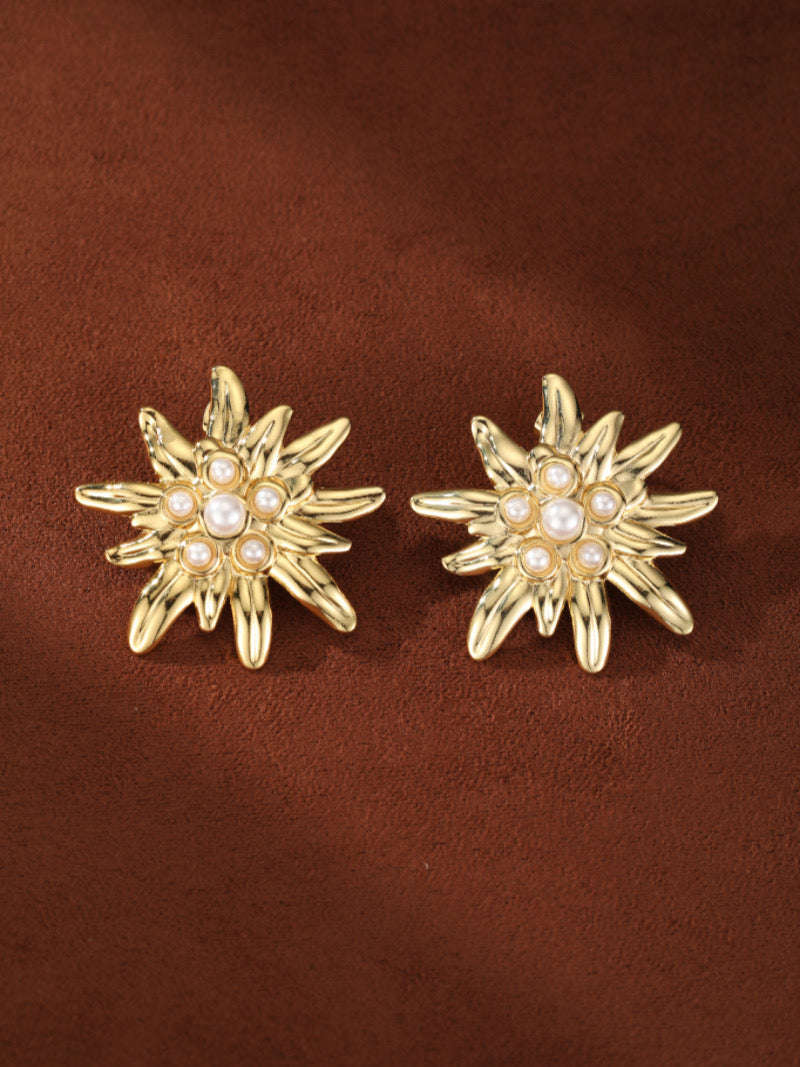 A pair of gold-toned, flower-shaped stud earrings with a central cluster of imitation pearls on a textured brown background.