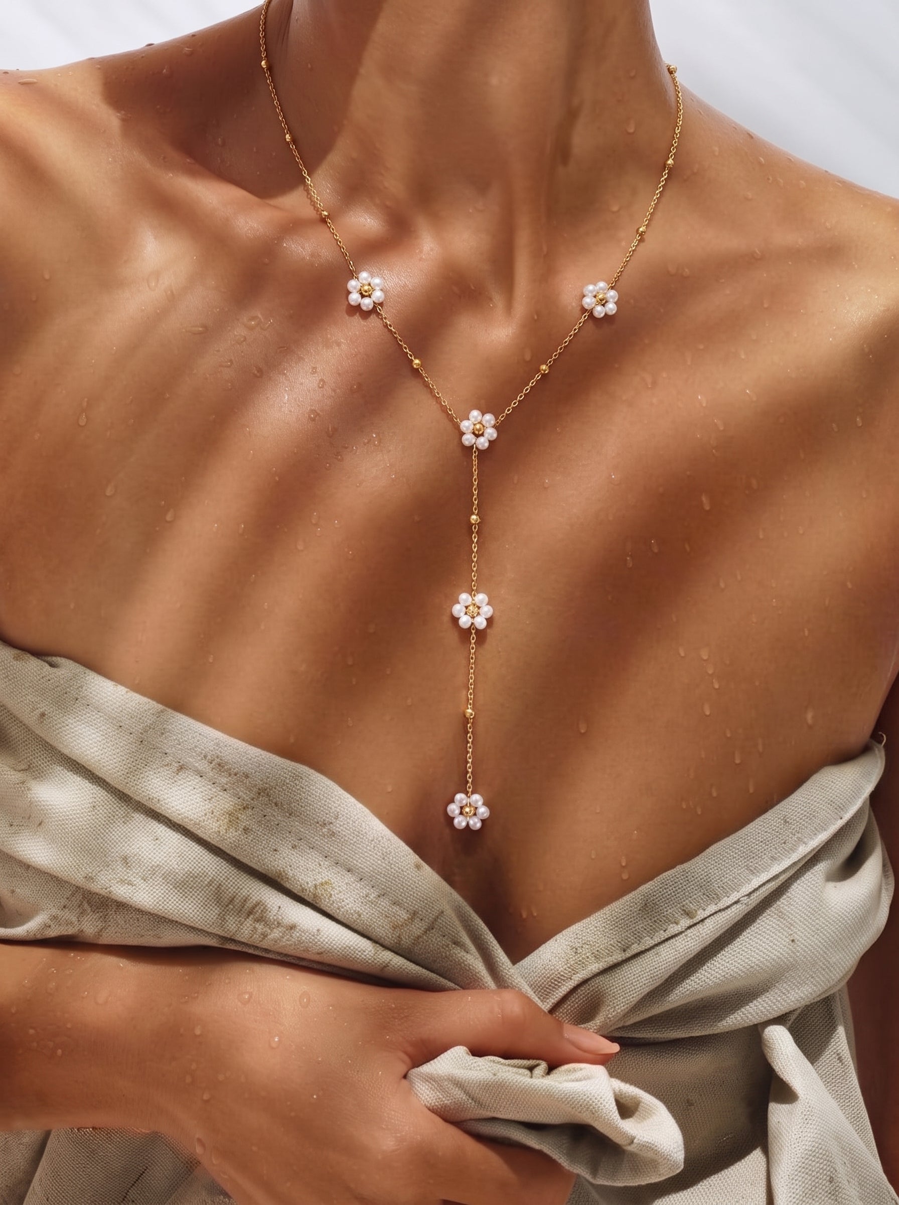 A delicate gold chain necklace with multiple flower charms, each featuring small white freshwater pearls and a gold bead center, rests on a woman's décolletage, suggesting a summer holiday vibe. @lifestyle