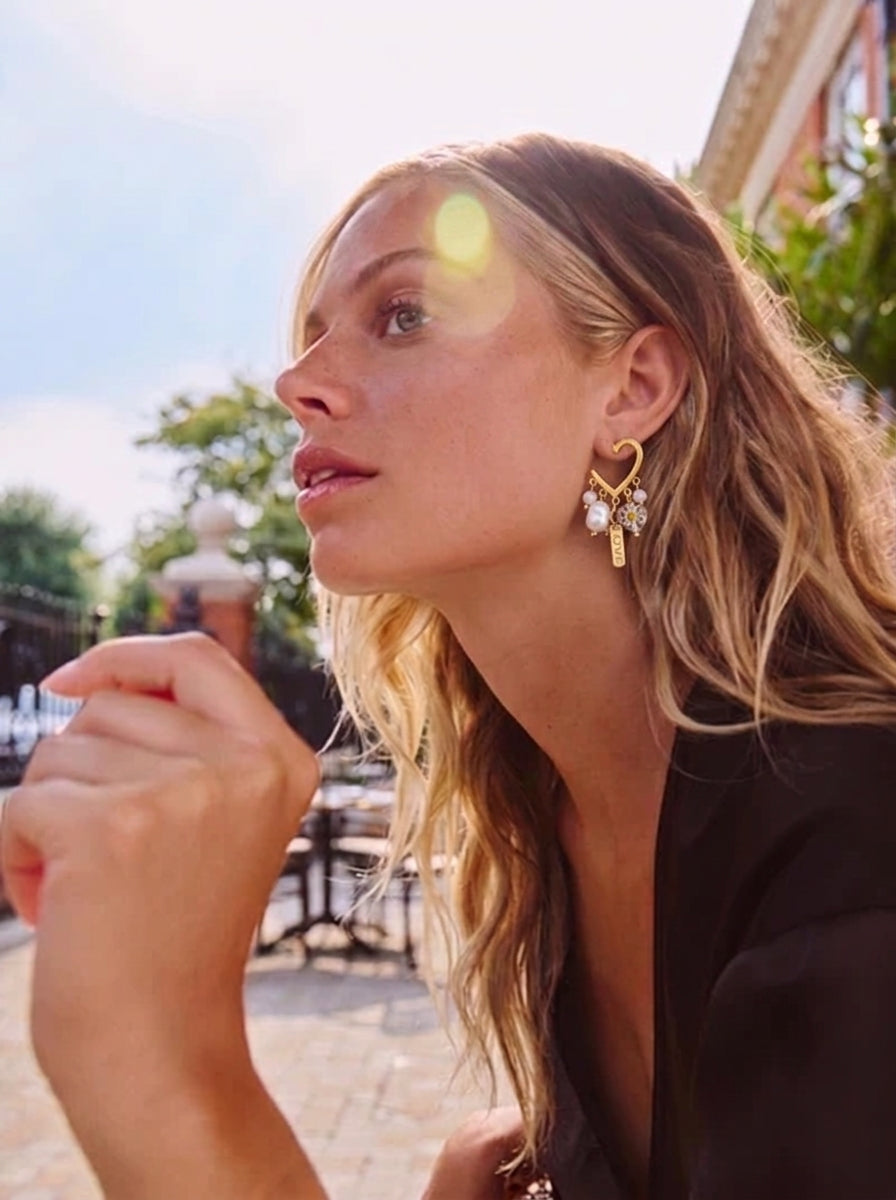 Une femme blonde portant des boucles d'oreilles pendantes en cœur doré avec perles d'eau douce et breloques, à l'extérieur dans un café. @lifestyle