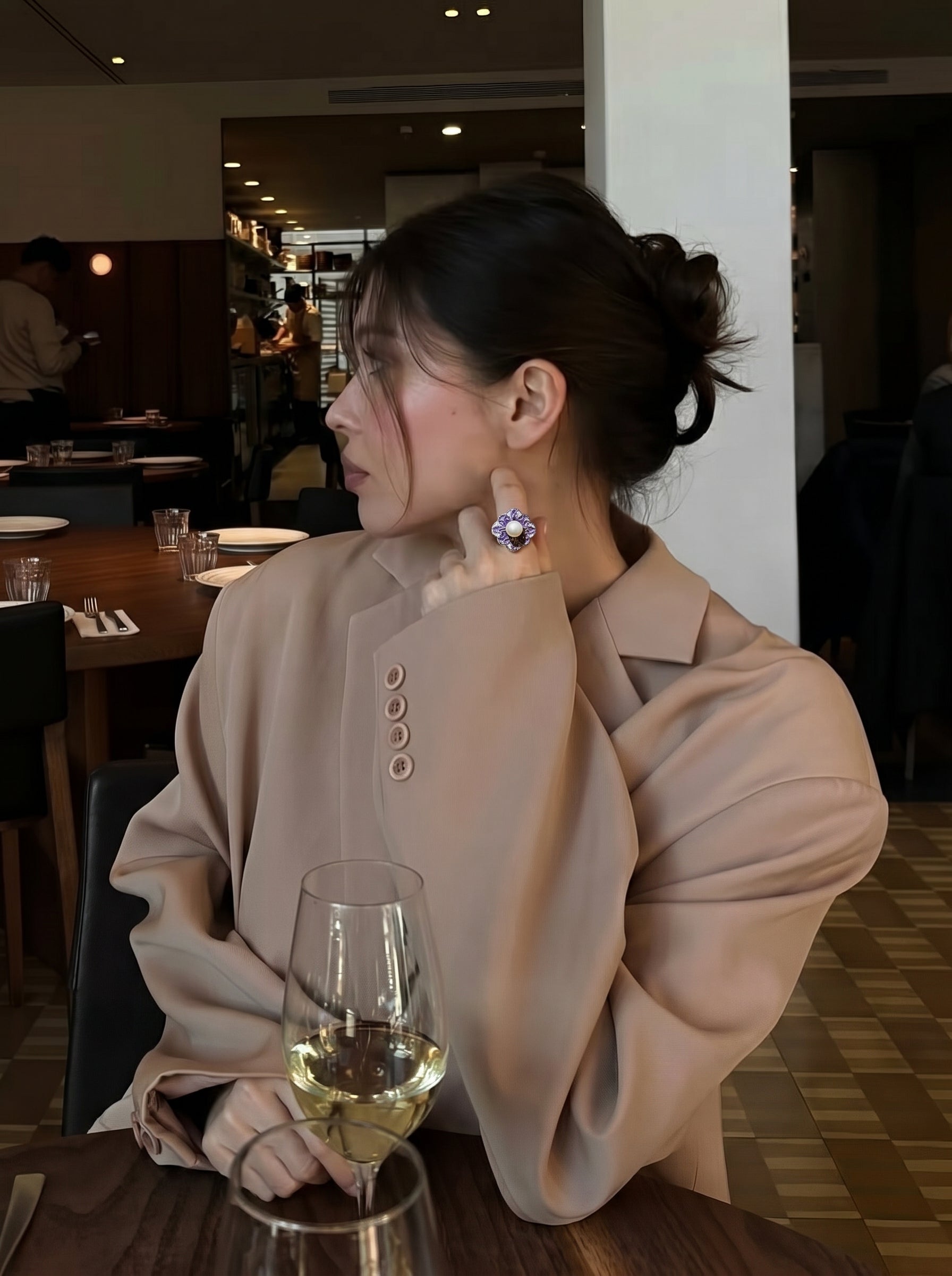 Une femme en blazer beige porte une bague manchette fleur Flora en perle d'eau douce aux pétales en émail violet et une perle blanche, assise à une table avec des verres de vin. @lifestyle