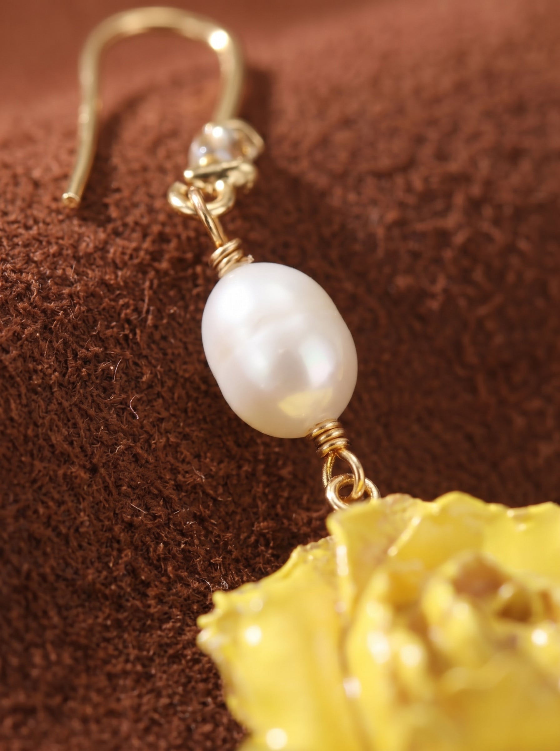 Gold dangle earrings featuring a white baroque freshwater pearl suspended above a bright yellow flower motif, set against a textured brown background. @pearl
