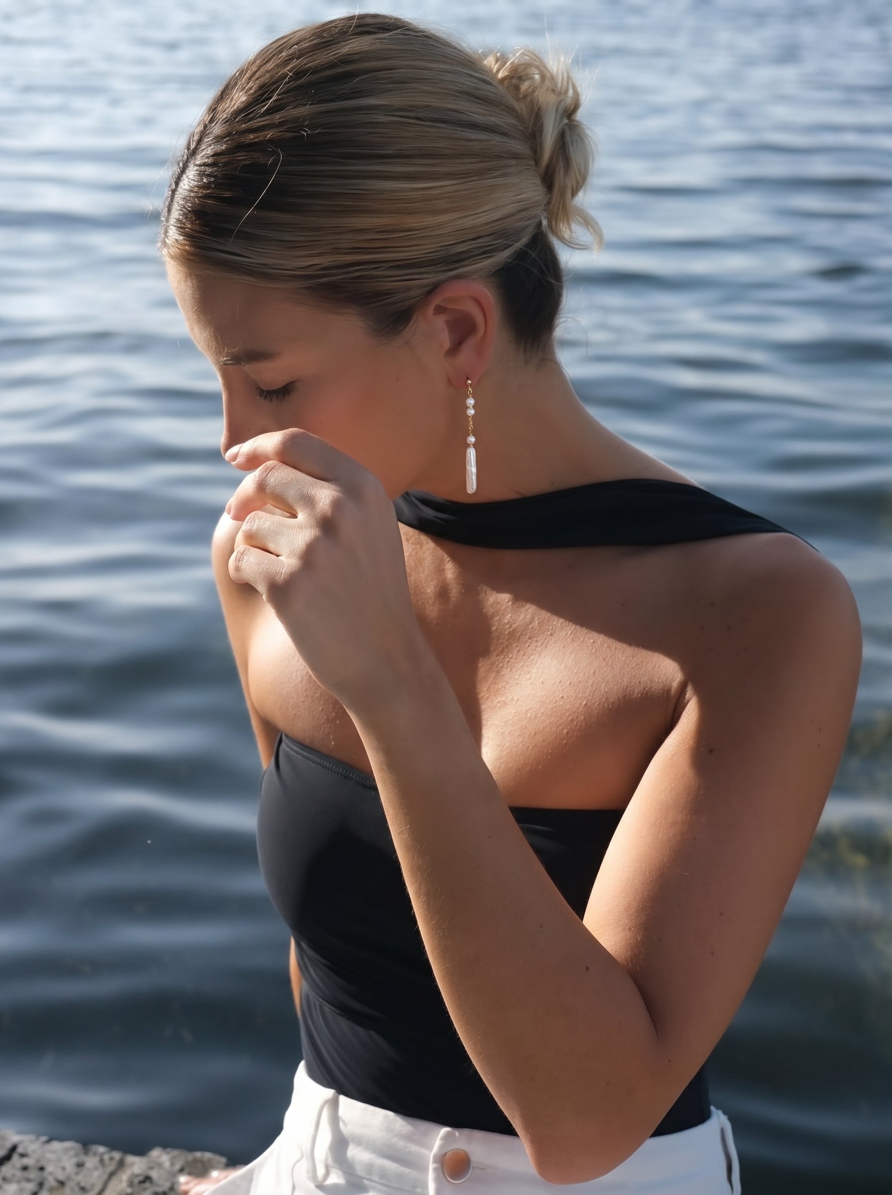 A woman with her hair in a bun wears a black strapless top and white pants, adorned with Flow Freshwater Pearl Long Dangle Earrings featuring small seed pearls and a long baroque pearl drop, by the water. @model