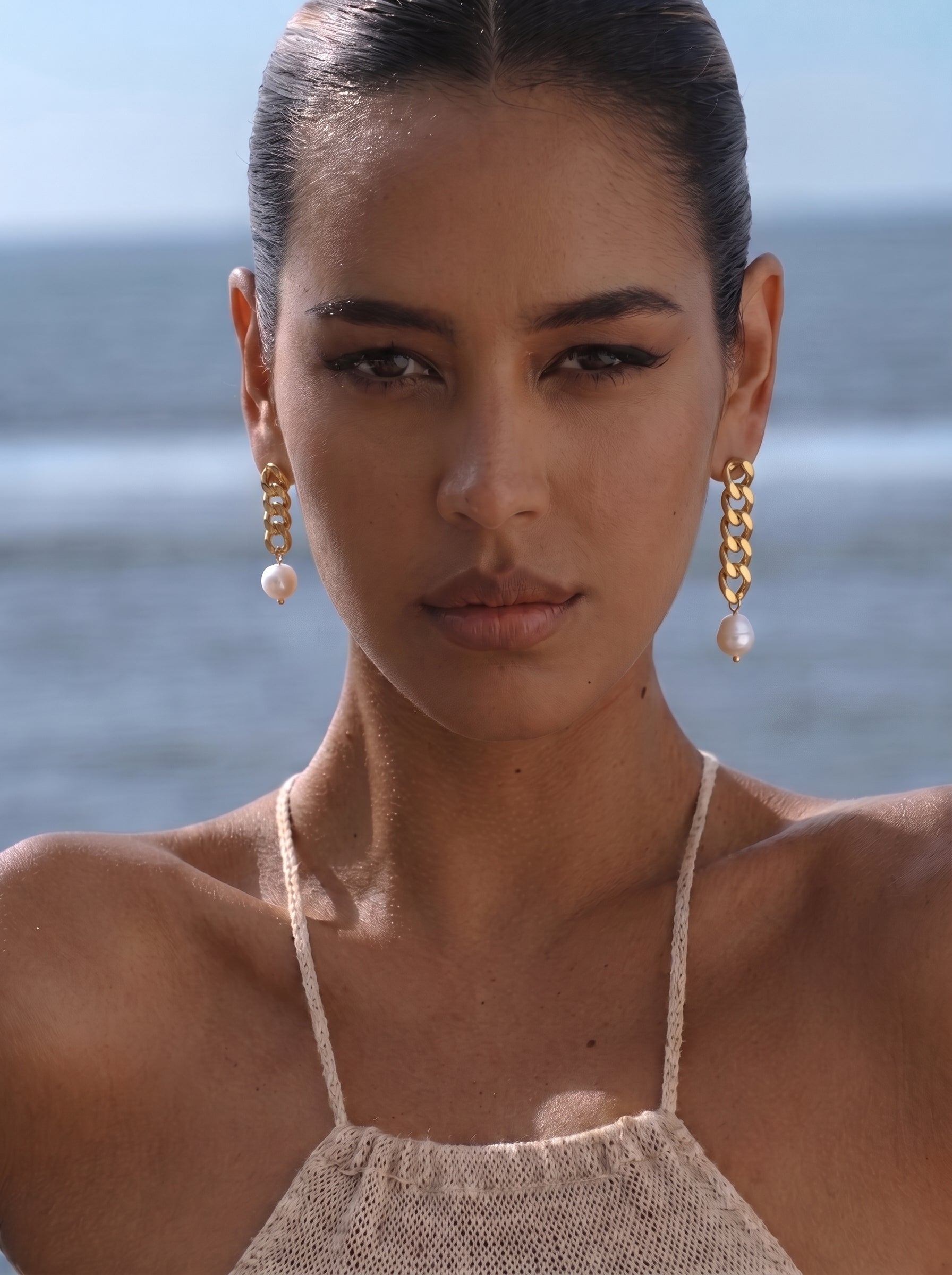 A woman with her face close up, wearing gold chain dangle earrings with white freshwater pearls, with the ocean blurred in the background. @lifestyle