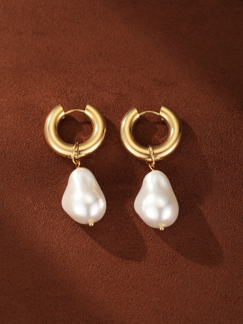 Gold-toned chunky hoop dangle earrings featuring irregular 10 mm freshwater white baroque pearls, on a brown textured background.