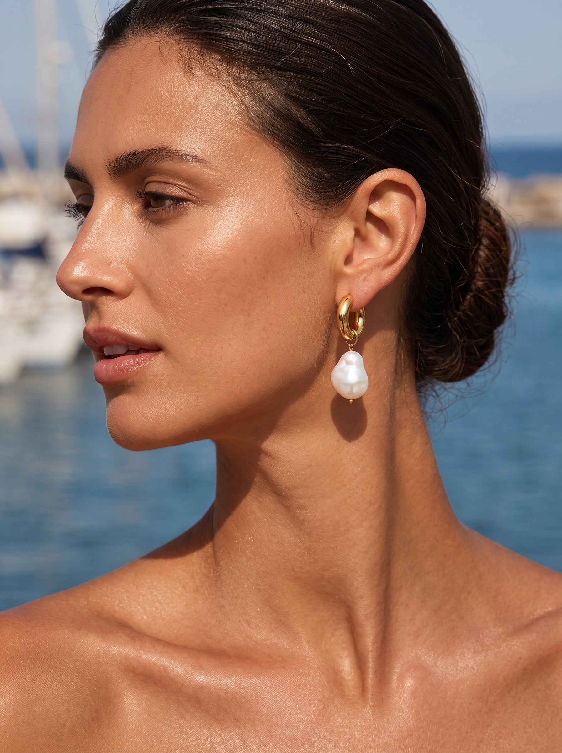 A woman with her hair pulled back wears the Forma Freshwater Pearl Chunky Hoop Dangle Earrings, with a large baroque pearl hanging from a gold hoop, against a backdrop of blue water and boats. @lifestyle