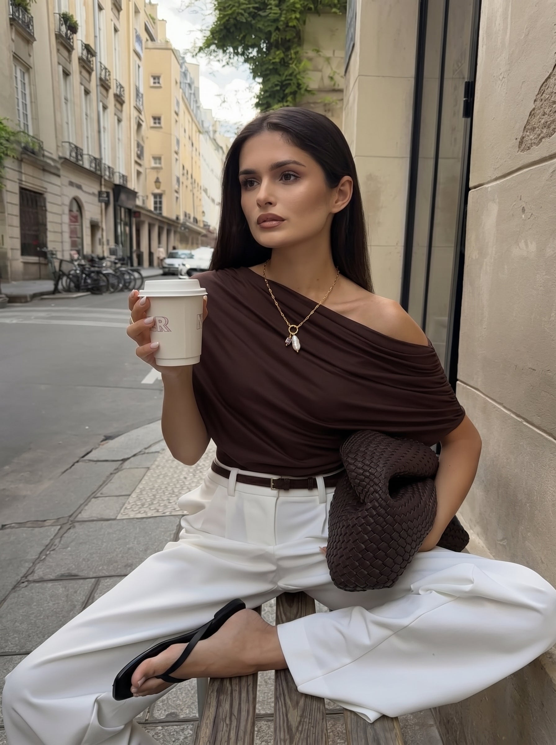 Une femme en haut marron et pantalon blanc est assise à l'extérieur sur un banc, tenant une tasse de café et portant le collier pendentif gemme Gaiety en perle d'eau douce avec un pendentif en perle baroque. @lifestyle