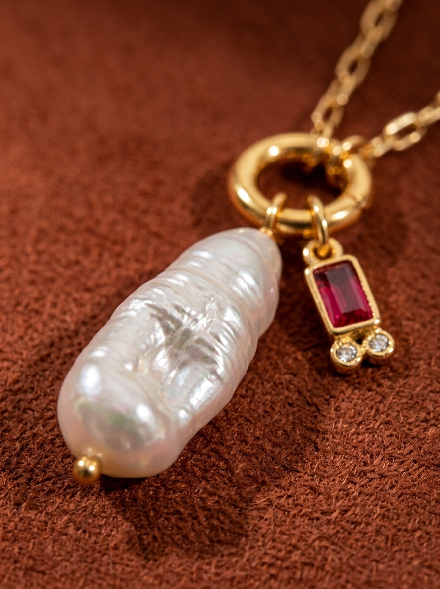A close-up shot of a Gaiety necklace featuring a large, elongated baroque freshwater pearl and a small rectangular red gemstone with two small clear stones, all set in gold-tone metal on a textured brown background. @pearl