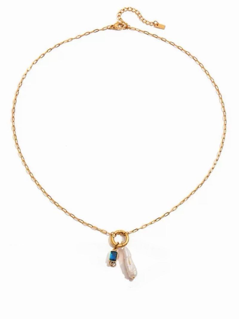 A gold-tone necklace with a delicate chain, featuring a circular pendant. Dangling from the pendant are a rectangular blue gemstone with pavé accents and an elongated Baroque freshwater pearl.