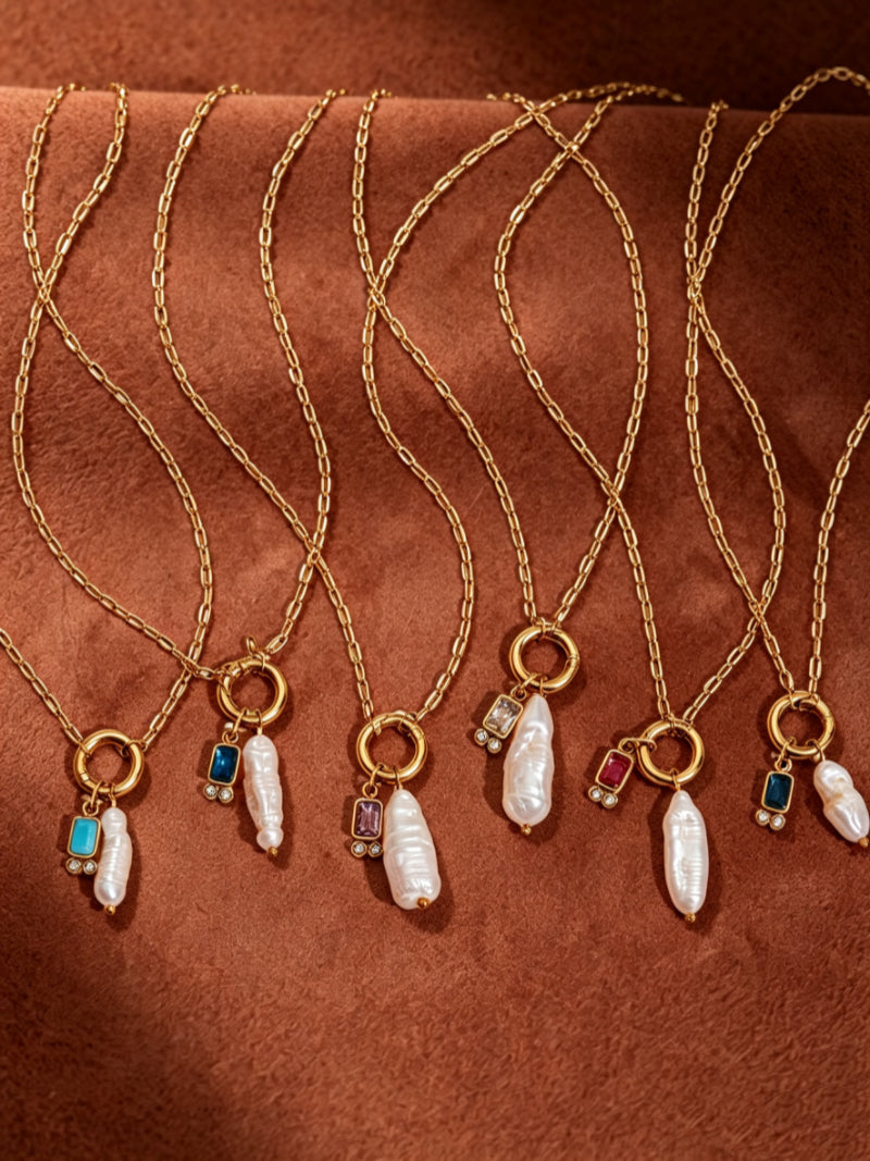 A collection of seven gold-tone necklaces, each featuring a unique baroque freshwater pearl drop accented with colorful gemstones like turquoise, amethyst, ruby, and sapphire, set against a textured brown background.