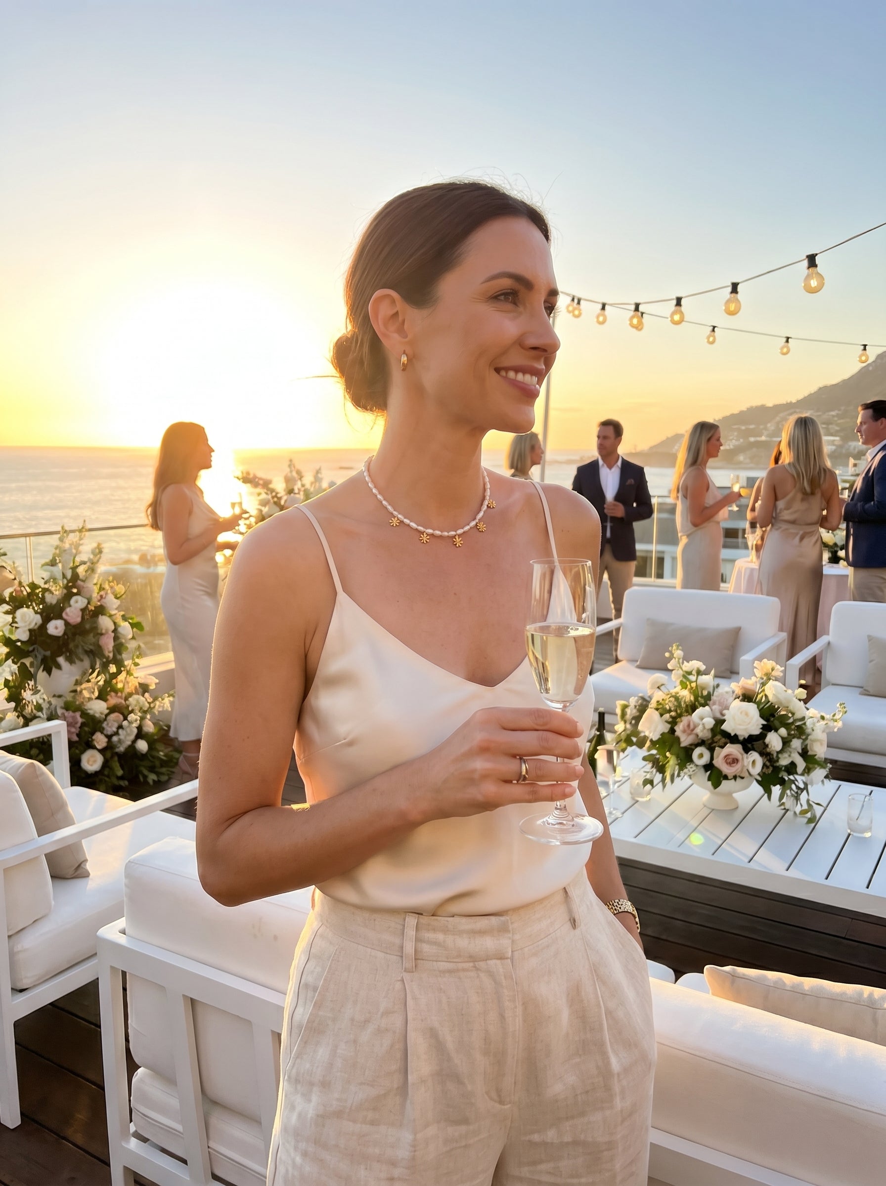 A woman wearing a white freshwater pearl choker necklace with golden flower charms, holding a champagne flute at a sunset outdoor party. @lifestyle #lifestyle