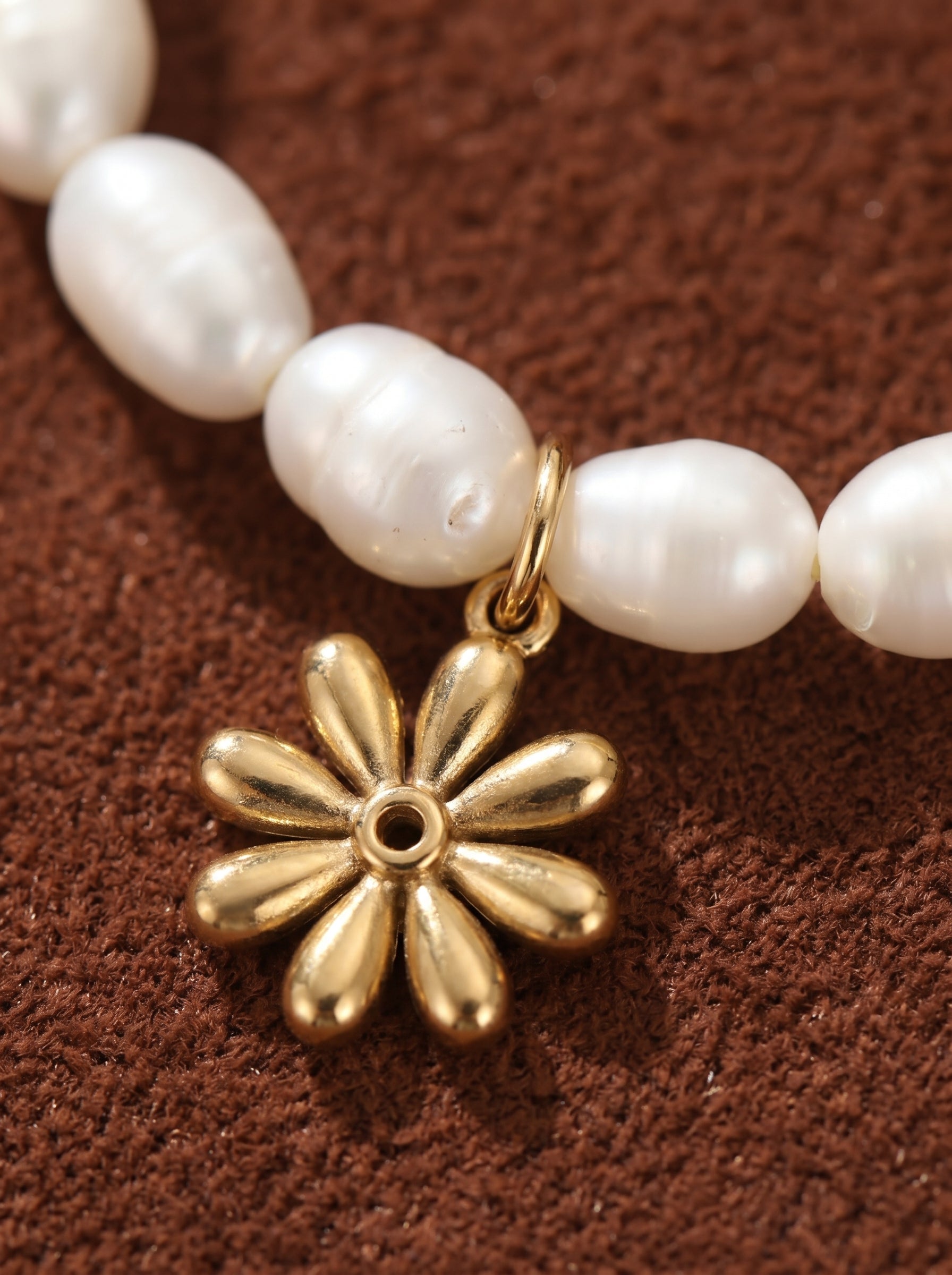 Close-up of a Gemma Strand choker necklace featuring lustrous white freshwater seed pearls and a small, golden flower charm. The pearls are strung together on a fine chain, with the golden flower charm adding a touch of whimsical elegance. The necklace rests on a textured brown surface. @pearl