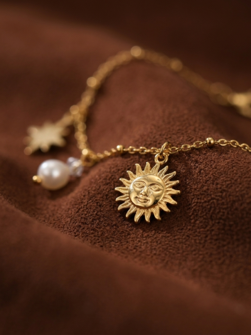 A close-up studio shot of the Glint bracelet, featuring a delicate gold chain with a sun charm, a star charm, and a baroque freshwater pearl. The bracelet is displayed on a rich brown, textured surface.