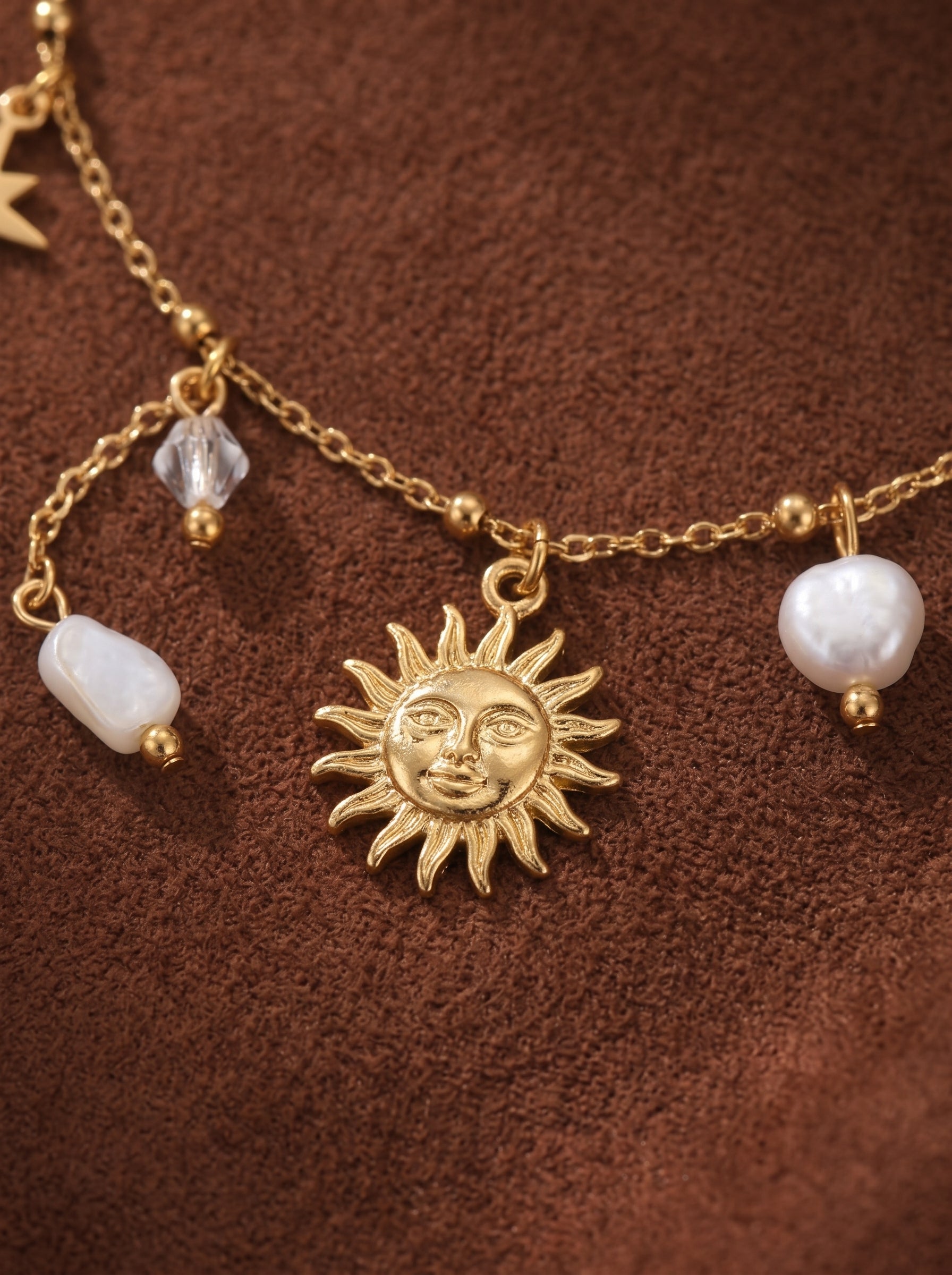 Close-up of a gold-tone chain bracelet featuring a sun charm, baroque freshwater pearls, and clear faceted beads on a brown textured background.