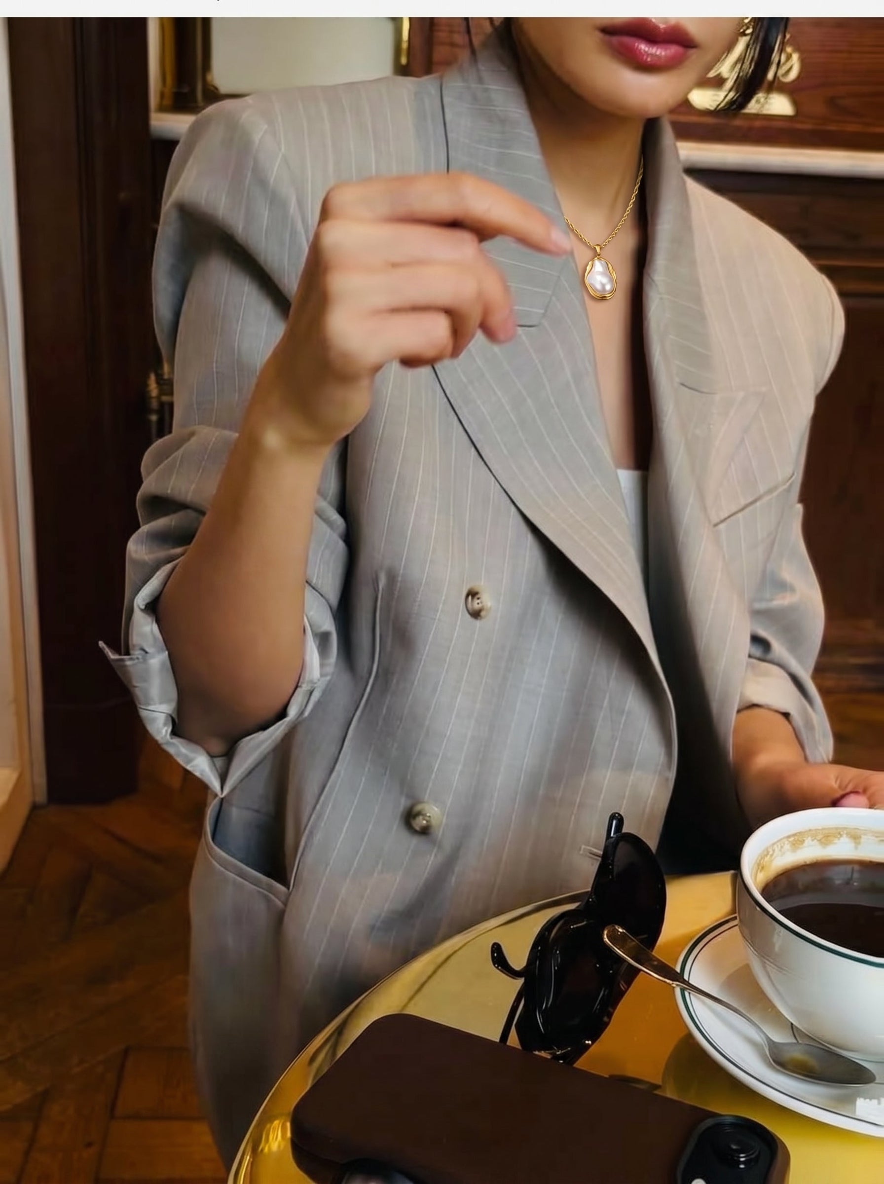 A woman wearing a pinstripe blazer and a chunky gold chain necklace with a large baroque freshwater pearl pendant, sitting at a table with coffee and sunglasses. @model #lifestyle