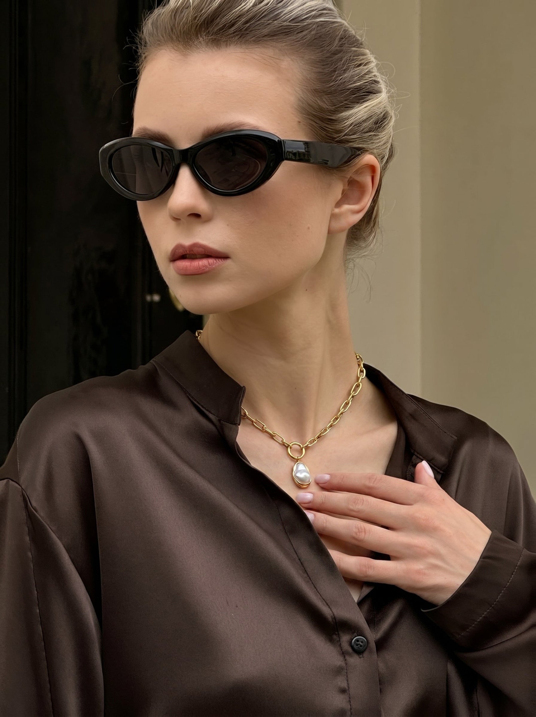 A woman wearing a brown satin shirt and black sunglasses, showcasing the Golden Tide chunky gold chain necklace with a large, irregular white freshwater pearl pendant.#lifestyle