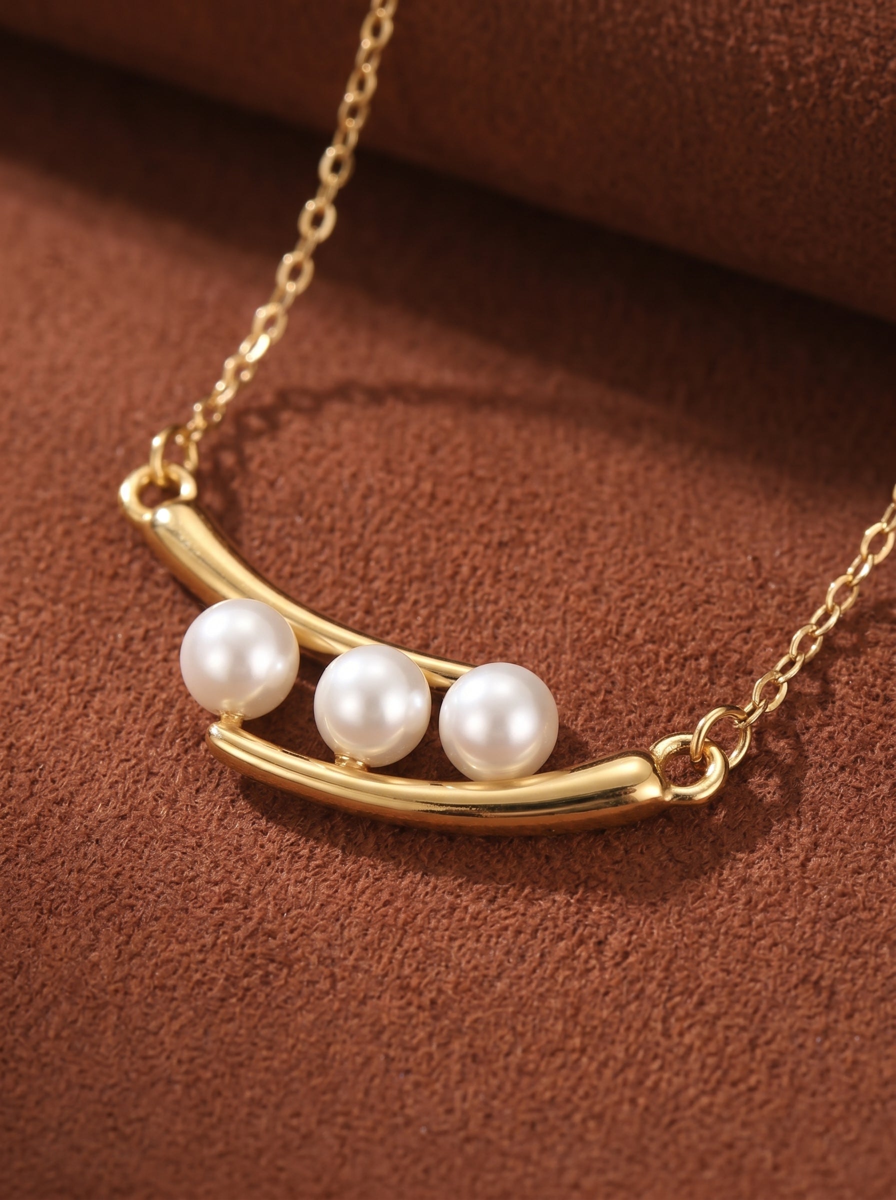 Gold-tone curved bar pendant necklace featuring three 4mm round white imitation pearls on a delicate chain, presented against a brown textured background.@pearl