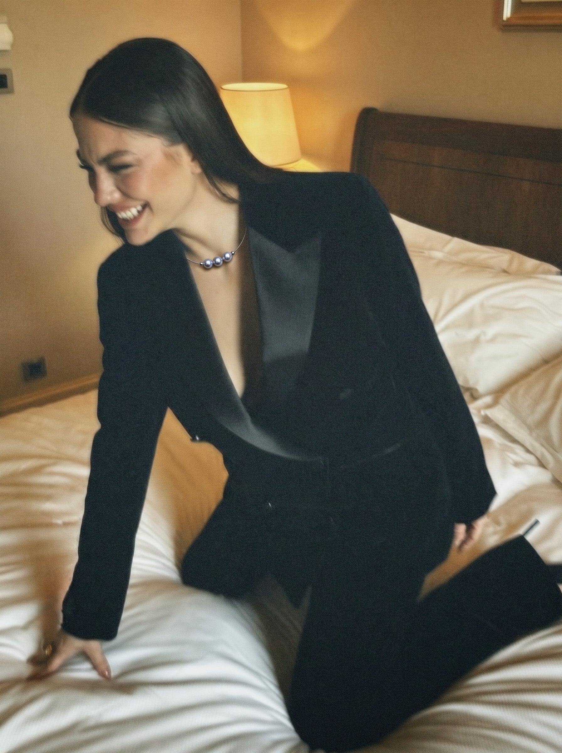 A smiling woman wearing a black tuxedo and the Haze Akoya Pearl Floating Chain Necklace, sitting on a bed. The necklace features three luminous Akoya pearls on a delicate chain. @lifestyle