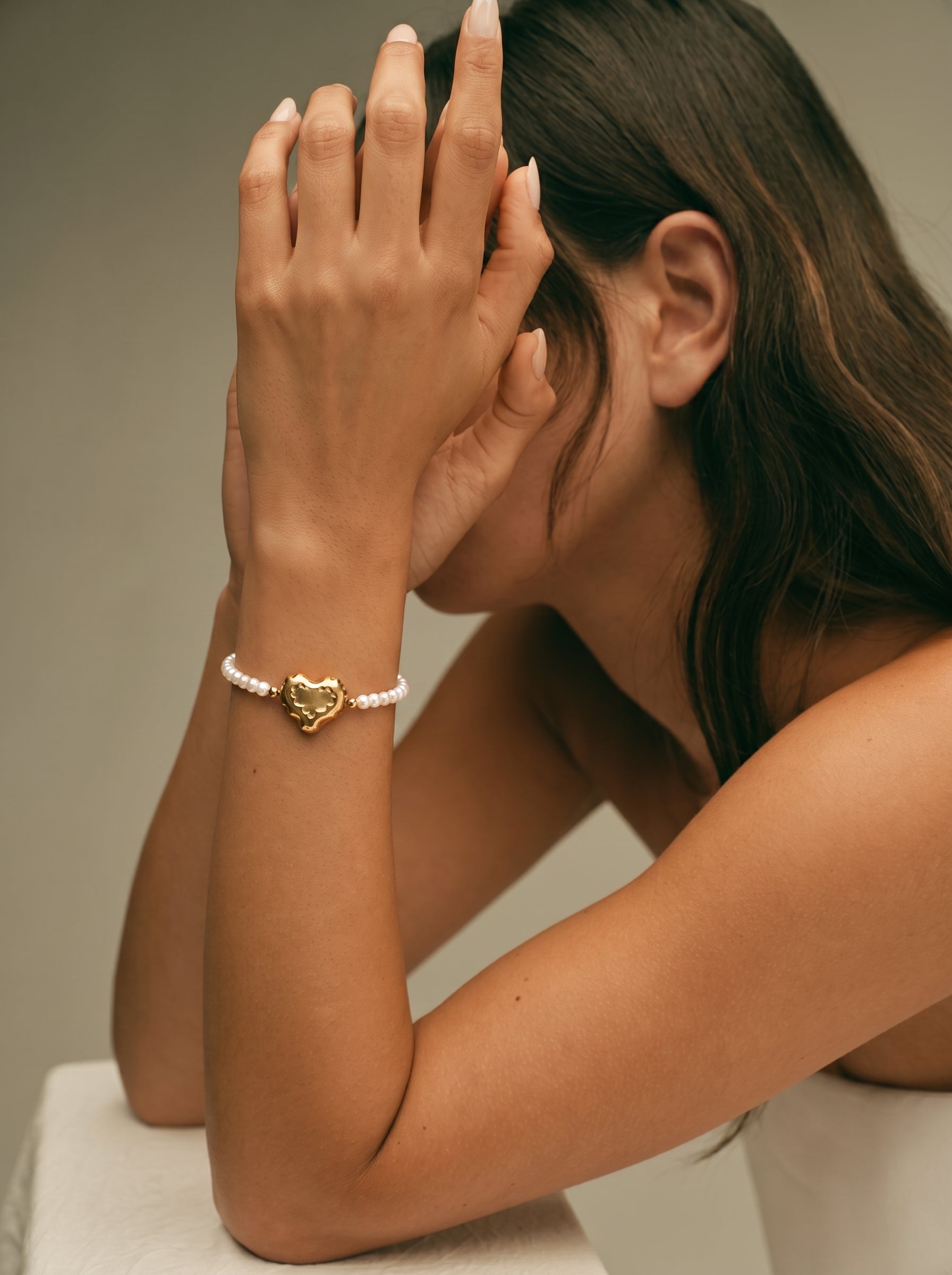 A woman's arm wearing a delicate bracelet made of small white freshwater pearls and a central, slightly puffed gold-toned heart charm. @model