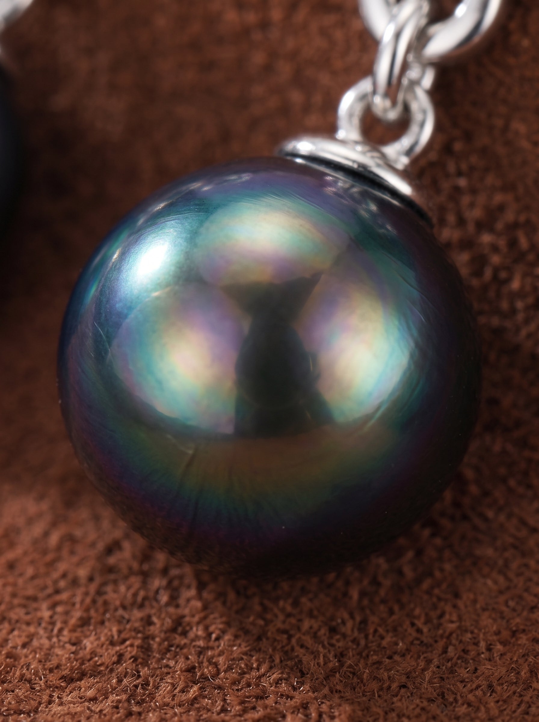 Close-up of a lustrous 10mm round Tahitian black pearl with iridescent hues, set in a silver drop earring with a stylized leaf motif. @pearl