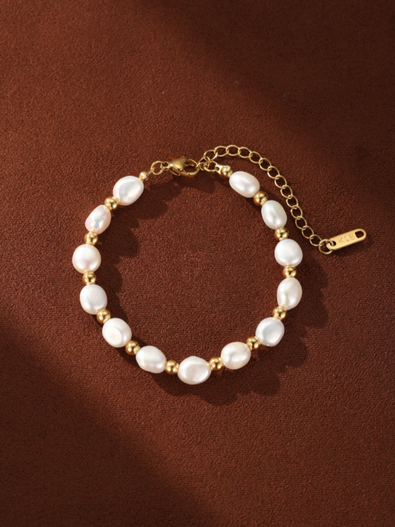 A gold-toned bracelet featuring irregular white freshwater Baroque pearls interspersed with small gold beads on a brown background.