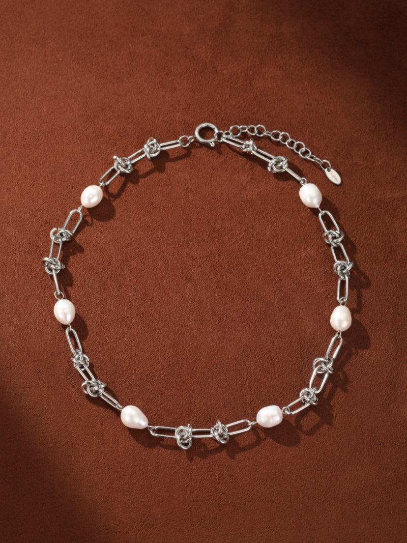 A silver choker necklace featuring a unique interlinked chain design with several lustrous white freshwater drop pearls spaced evenly. The necklace is displayed against a smooth brown background. @pearl