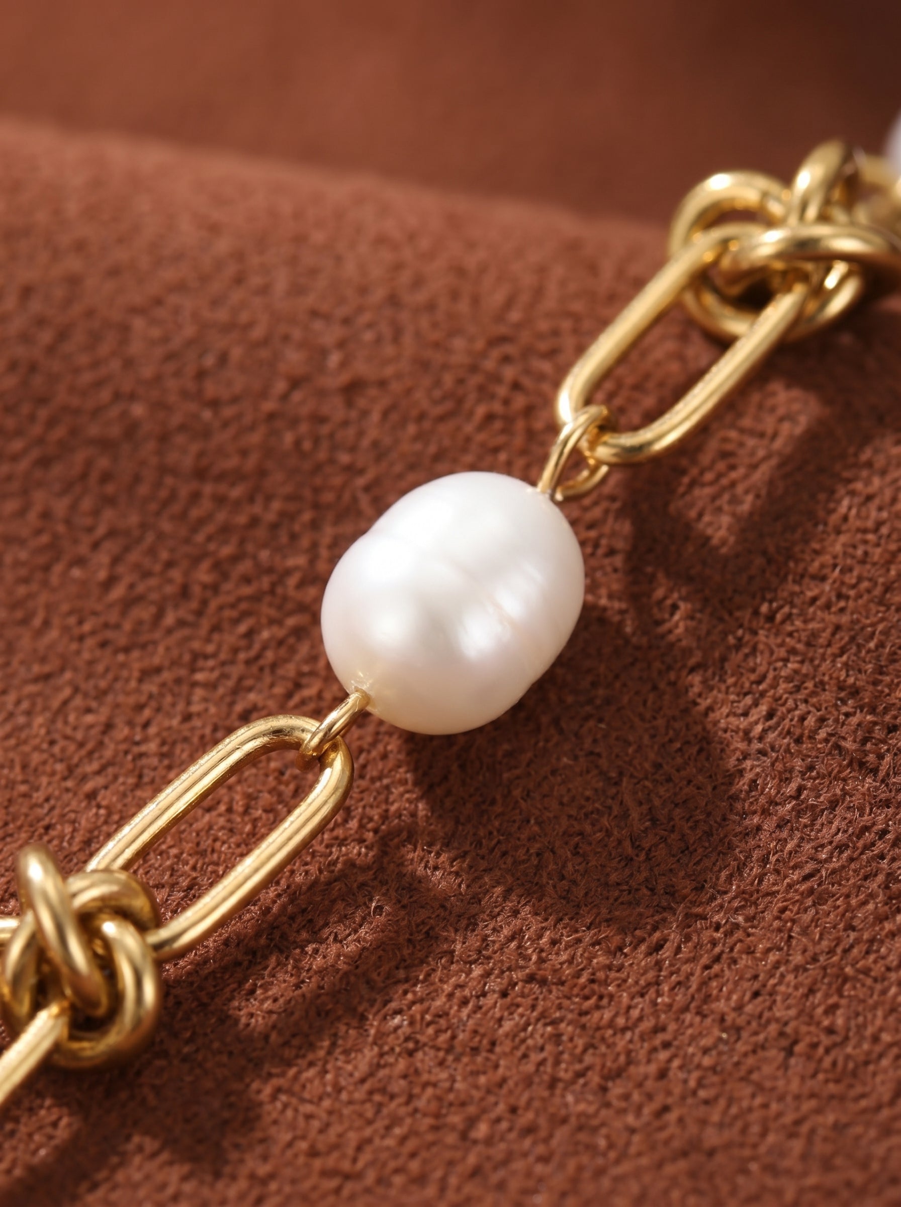 Close-up of a gold chain choker necklace featuring a single, lustrous white freshwater pearl. The chain has interlinked oval links with a twisted knot detail. The pearl is oval-shaped and has a natural, irregular beauty. The background is a textured brown surface. @pearl