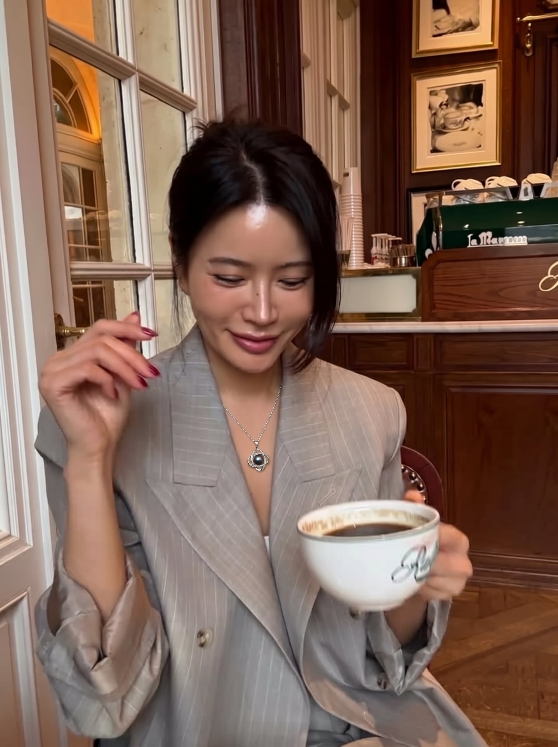 Une femme en blazer rayé gris tient une tasse de café, révélant le collier pendentif à chaîne quatrefeuille Interstellar en perle de Tahiti avec une perle noire et des détails en zircone cubique. @lifestyle