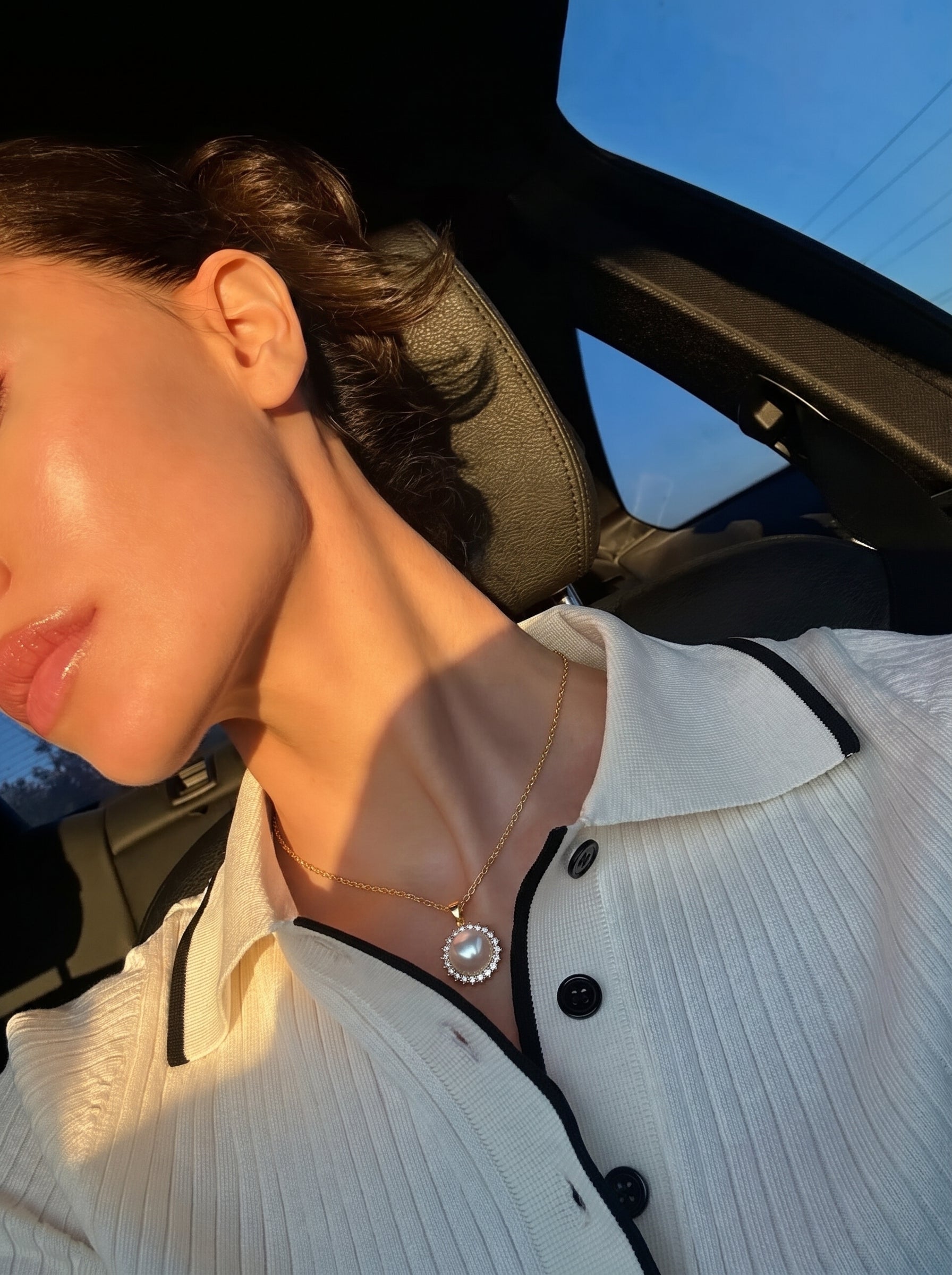 Gros plan d'une femme portant le collier pendentif soleil Iris en perle Akoya, une perle ronde avec un halo de pierres serties en or, avec la lumière du soleil illuminant son visage et le collier alors qu'elle est dans une voiture. @lifestyle