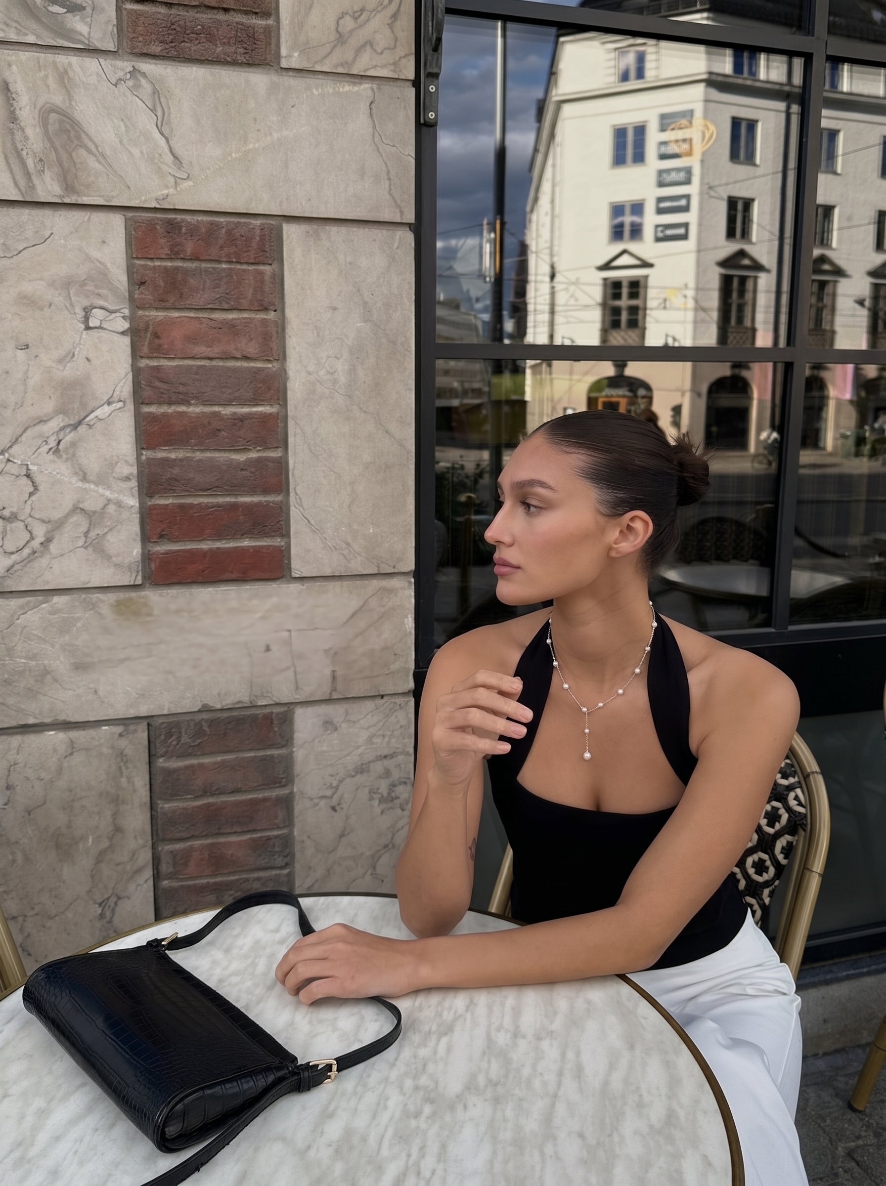 Une femme portant un haut licou noir est assise à une table de café en extérieur, accessoirisée du long collier à chaîne Journey en perle imitation. Le collier présente une chaîne délicate avec des perles imitation blanches alternées de 8 mm et 4 mm, et un pendentif en perle unique. Un sac à main à l'épaule embossé crocodile noir est posé sur la table à côté d'elle. @lifestyle