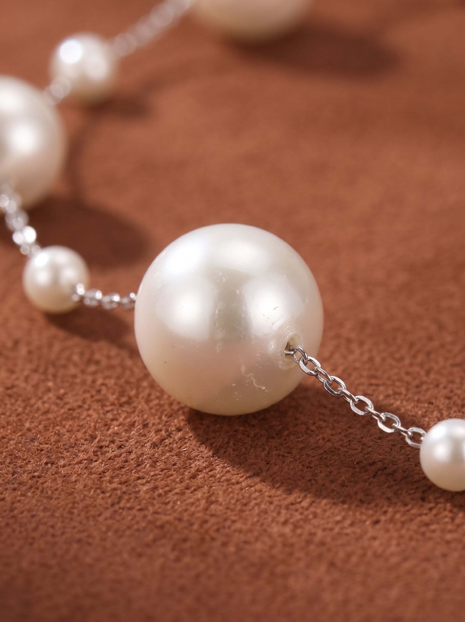 Close-up of a long chain necklace with alternating large and small white imitation pearls on a textured brown background. The delicate silver chain connects the lustrous pearls. @pearl