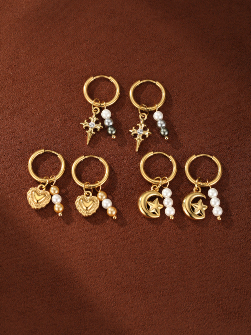 A collection of three pairs of gold hoop earrings, each with a different charm: a cross with pearls, a heart with pearls, and a moon with pearls.