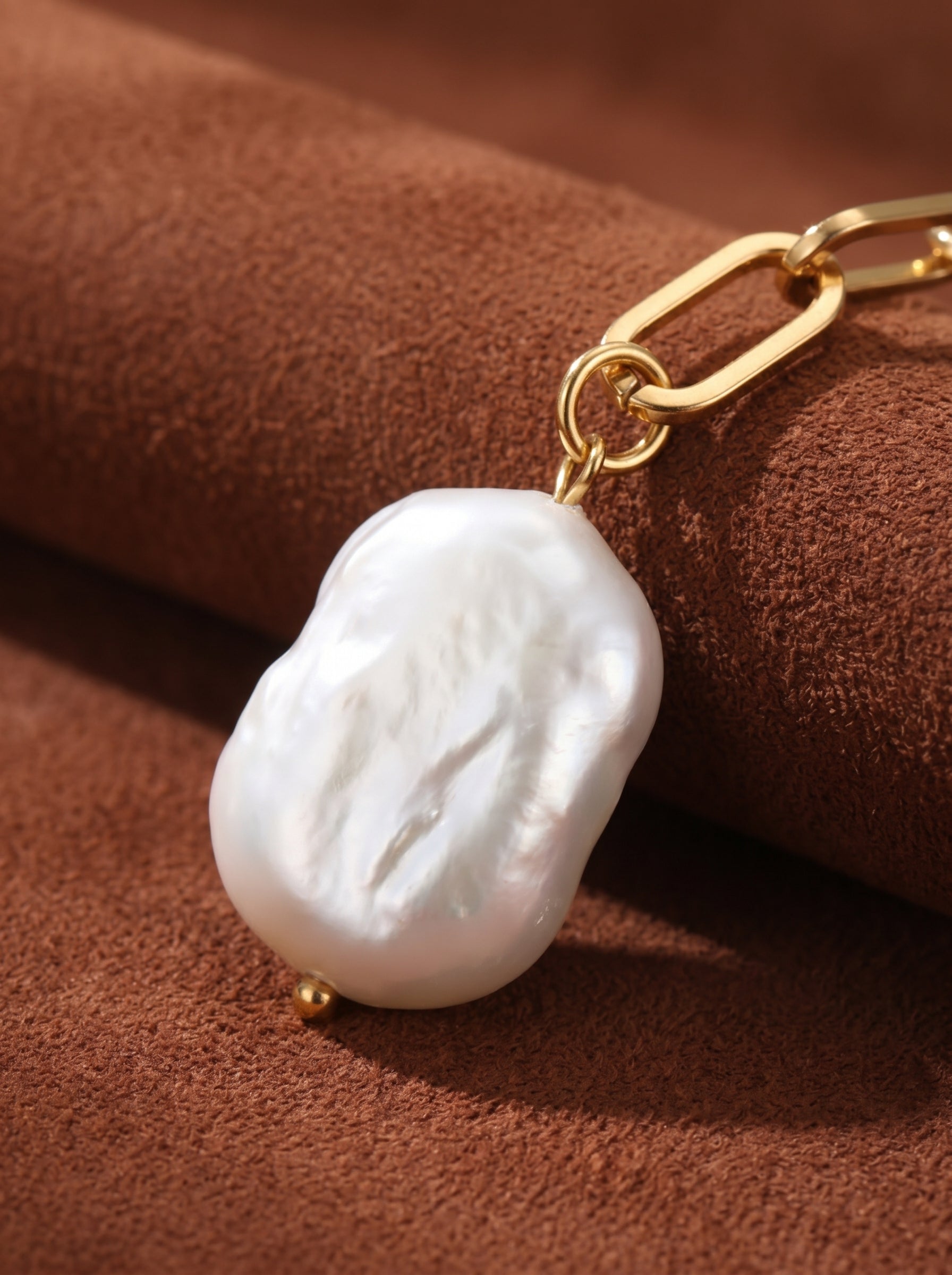 Close-up of a large, irregular white baroque freshwater pearl with a gold accent, attached to a chunky gold chain link necklace. The pearl has a luminous sheen and textured surface, set against a warm brown, suede-like background.@pearl