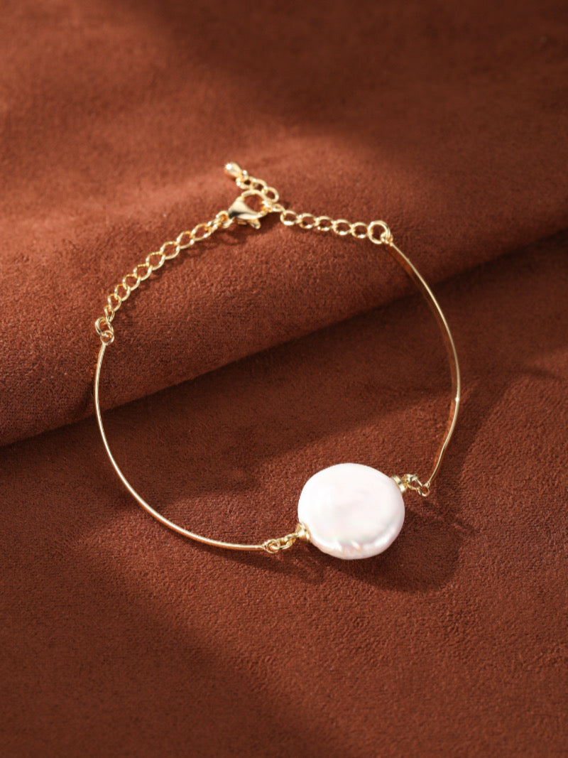 Gold-toned chain bracelet featuring a single, flat white freshwater pearl suspended on a thin bar, on a brown textured background.