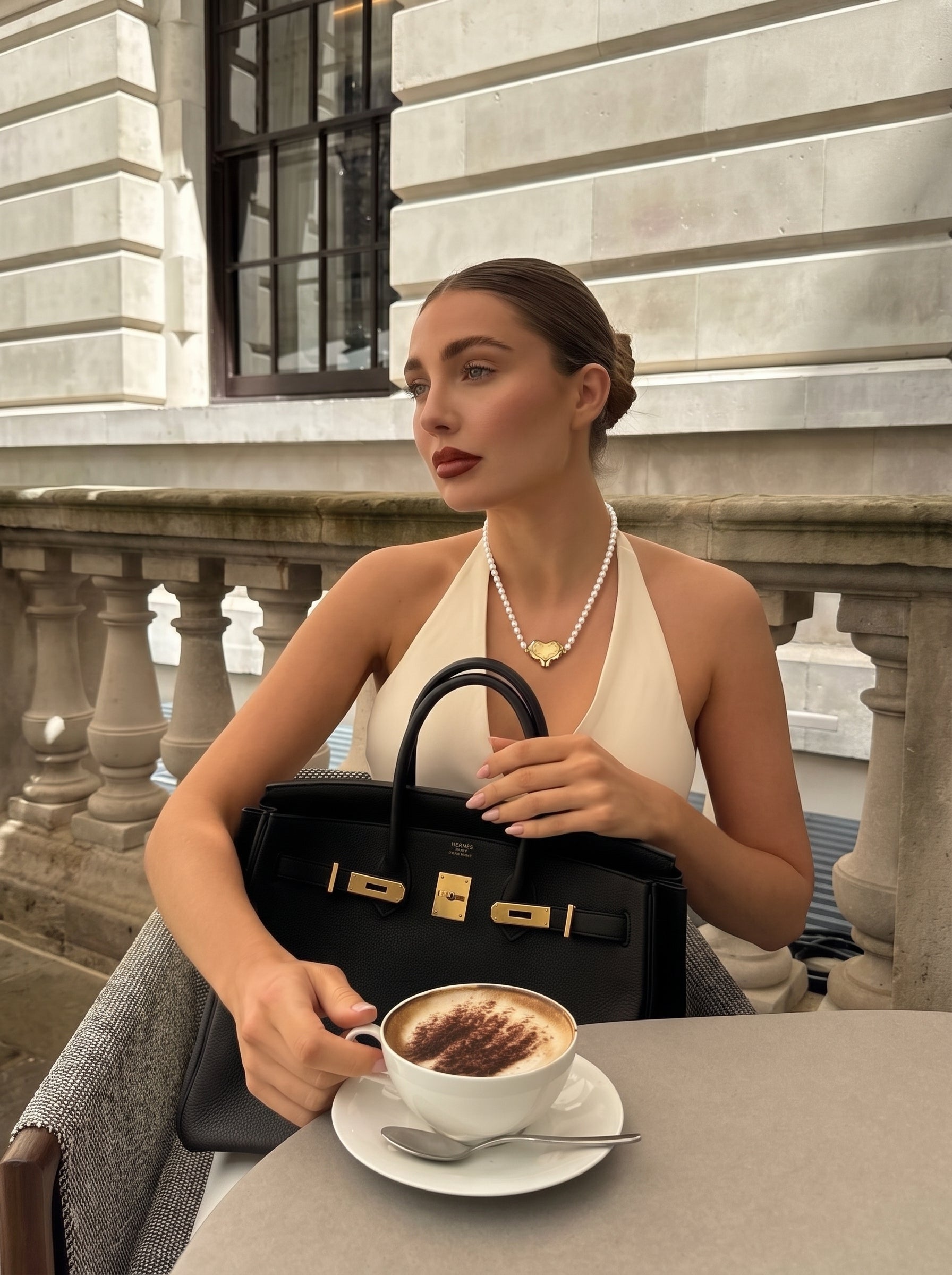 A woman wearing a white halter top and the Kindred Freshwater Pearl Heart Beaded Pendant Necklace, holding a black handbag and a cappuccino at a cafe table. @lifestyle