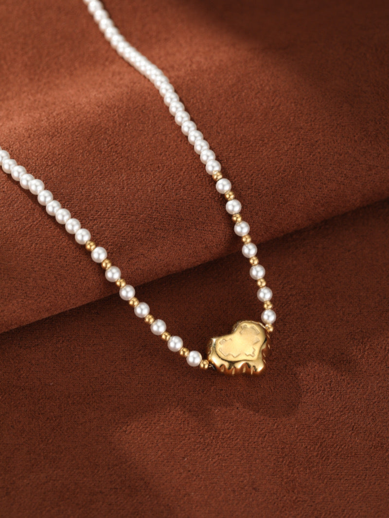 A close-up of the Kindred Freshwater Pearl Heart Beaded Pendant Necklace featuring a delicate strand of 3mm white pearls interspersed with gold beads, centered by a textured gold-tone heart pendant, against a brown background.