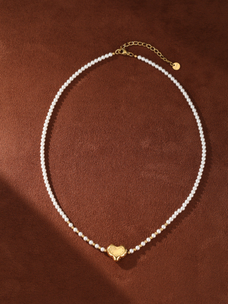 A delicate gold-tone necklace featuring a strand of small, luminous white freshwater pearls. A sculpted gold heart pendant is centered on the strand, accented by tiny gold beads. The necklace has an adjustable gold chain clasp.