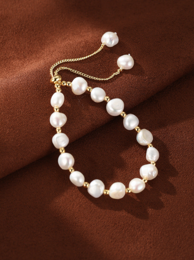Kinetic Strand bracelet featuring white freshwater pearls and gold-toned beads with an adjustable slider closure on a brown textured background.