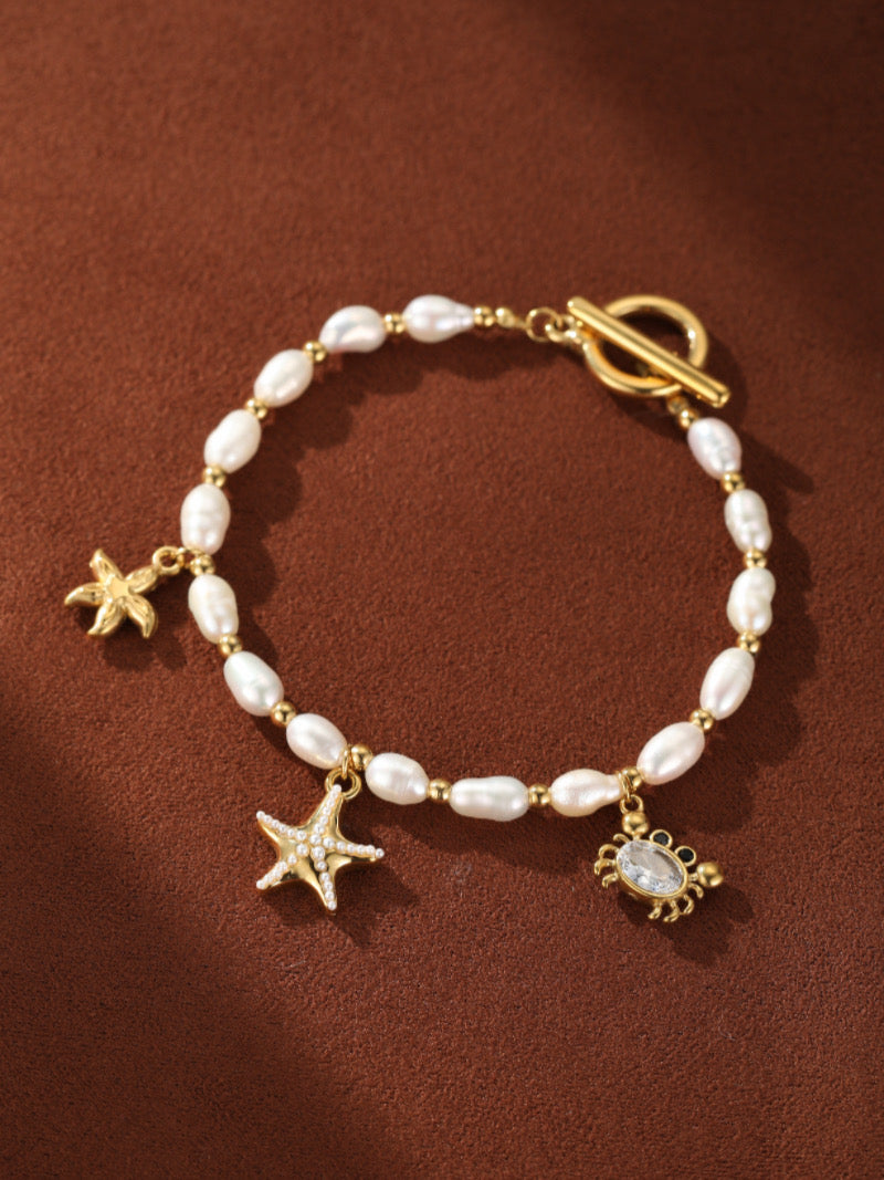 Lagoon bracelet featuring white freshwater seed pearls and gold oceanic charms including a starfish, whale, and a crab with a clear crystal, on a brown background.