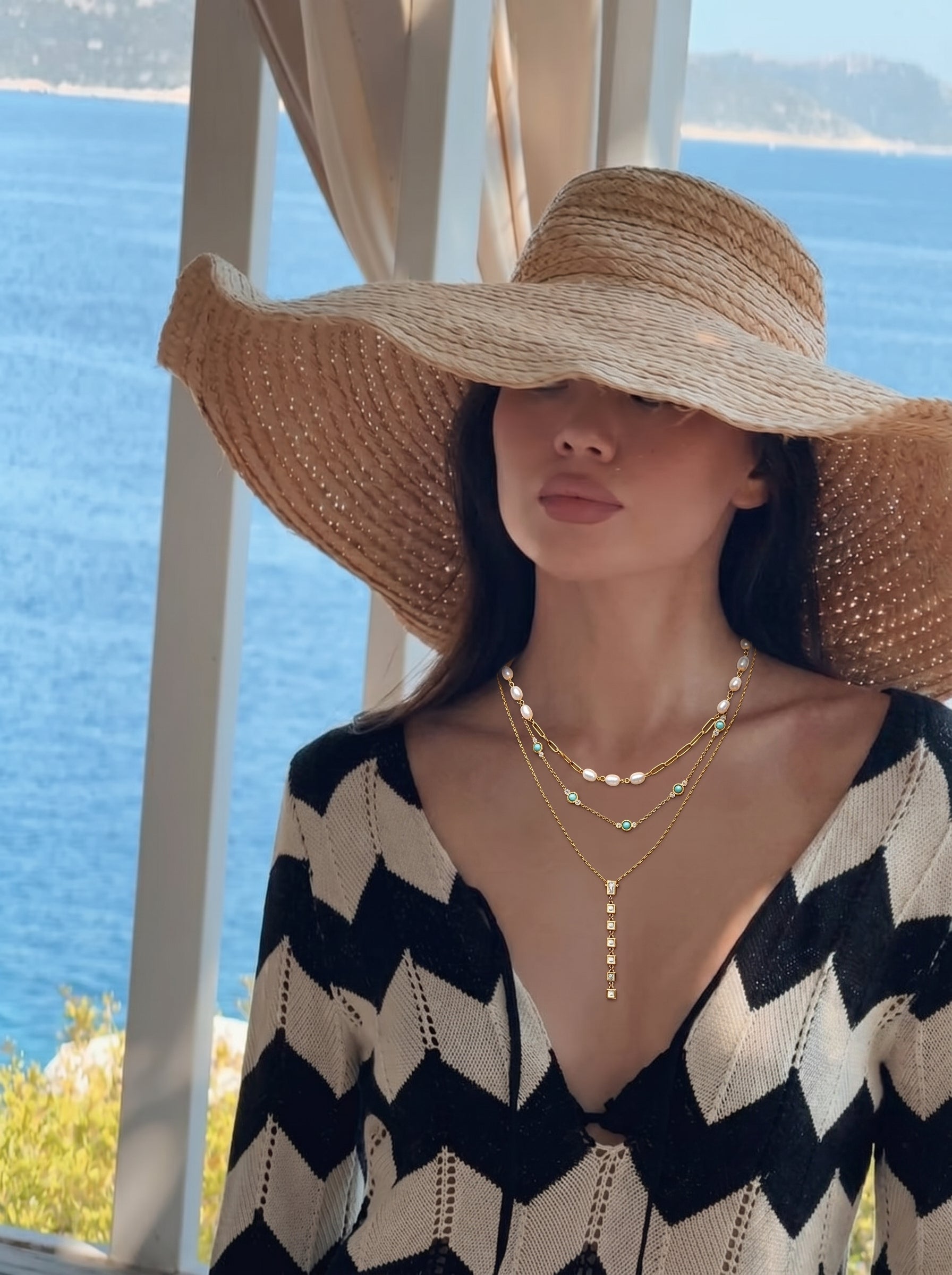 Une femme portant un chapeau de paille et un haut à motifs noir et blanc, parée du collier Lagoon Layers, présentant trois chaînes dorées avec des perles d'eau douce, des perles turquoise, et un pendentif goutte en pierres zircone cubique taille baguette et carrée. Le fond est une vue d'océan floue, indiquant un cadre lifestyle. @lifestyle