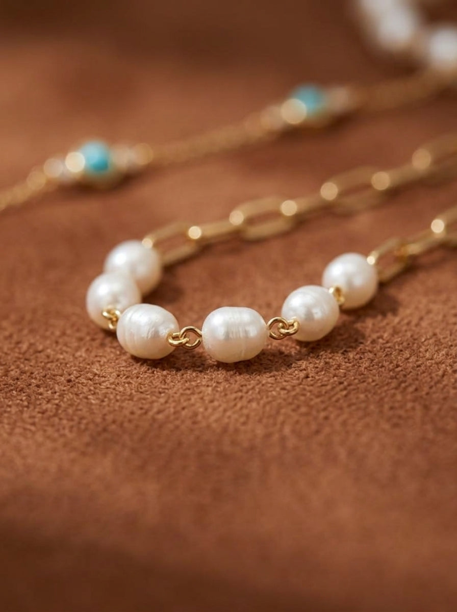 Close-up of a layered gold necklace featuring white freshwater seed pearls on the top chain and turquoise beads on a lower chain, with a textured brown background. @perle