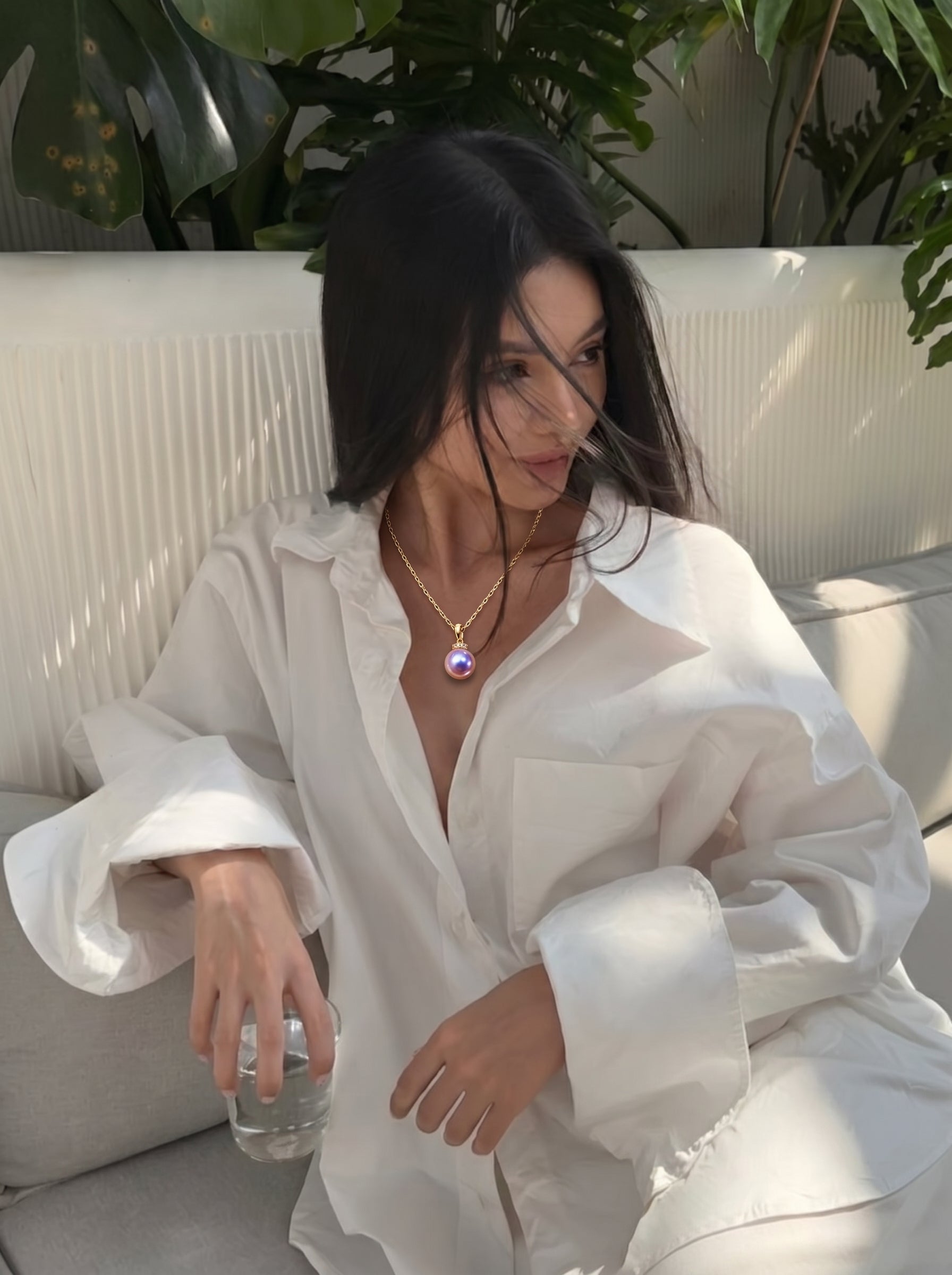 Une femme en chemise blanche oversize est assise sur un canapé, portant le collier Lila Drop avec un pendentif en perle violette d'eau douce, tenant un verre d'eau. @lifestyle