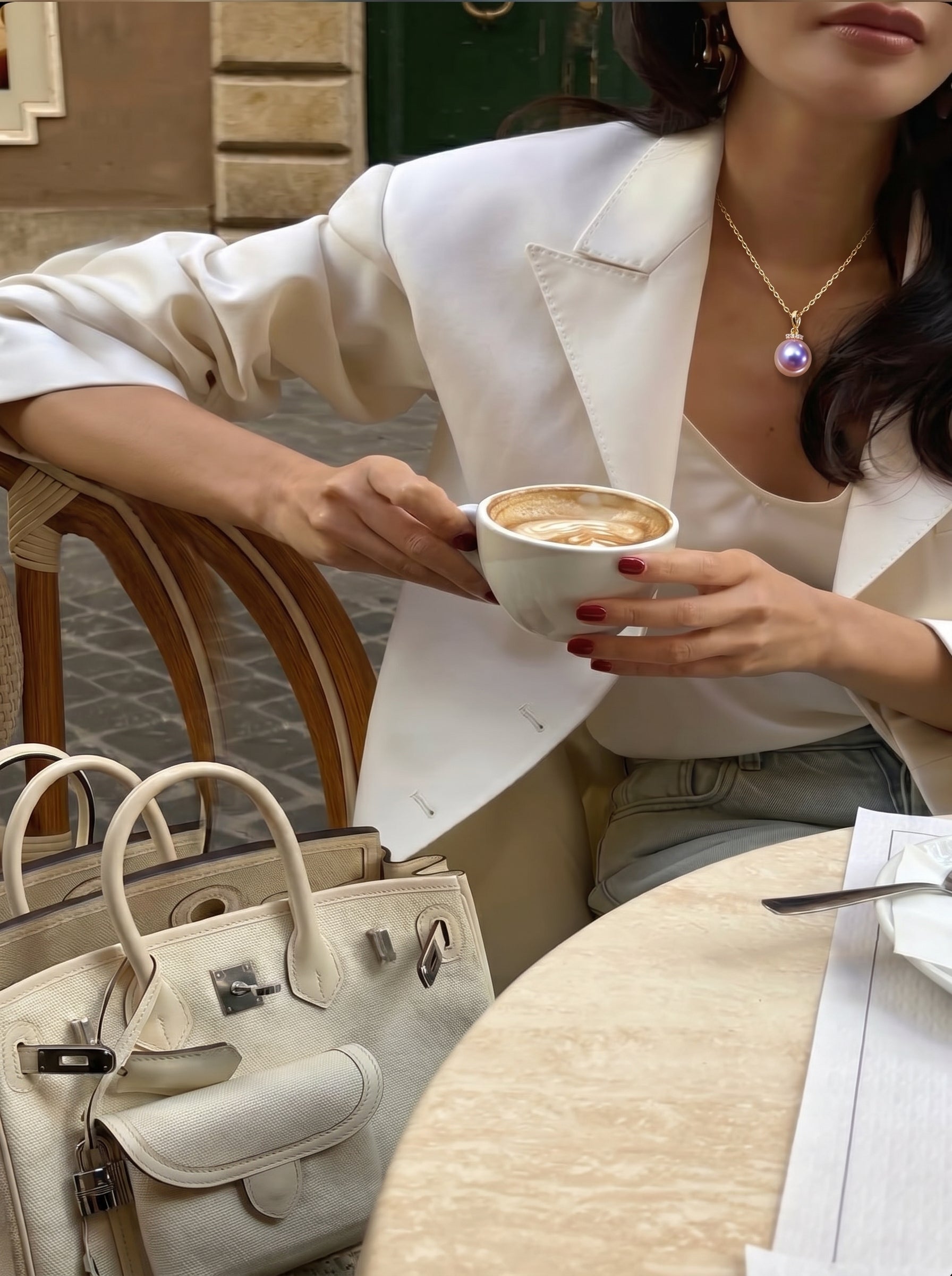 Femme en blazer blanc tenant une tasse de café, portant le collier pendentif à chaîne Lila Drop en perle d'eau douce avec une perle violette ronde et une bélière en cristal, dans un café en extérieur avec des sacs à main de créateur sur la table. @lifestyle