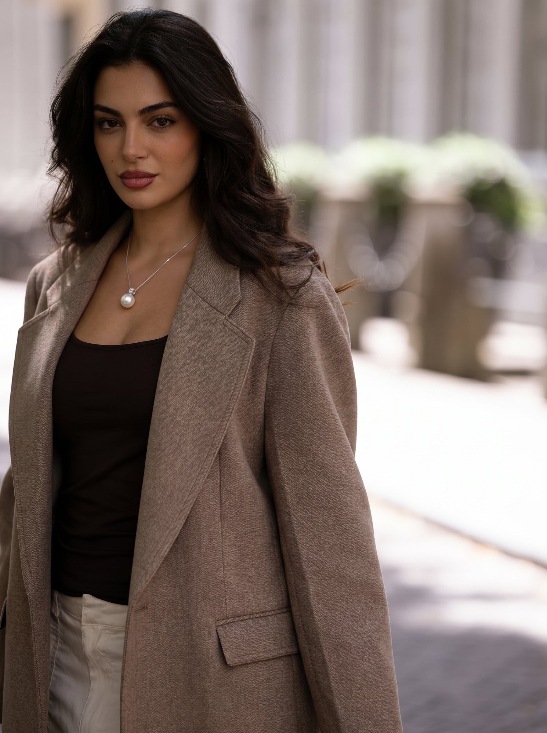 Une femme aux cheveux bruns porte un collier pendentif à chaîne Lumin en perle des mers du Sud présentant une grande et lustrée perle blanche sur une délicate chaîne argentée, superposé sur un blazer marron et un haut foncé. @lifestyle