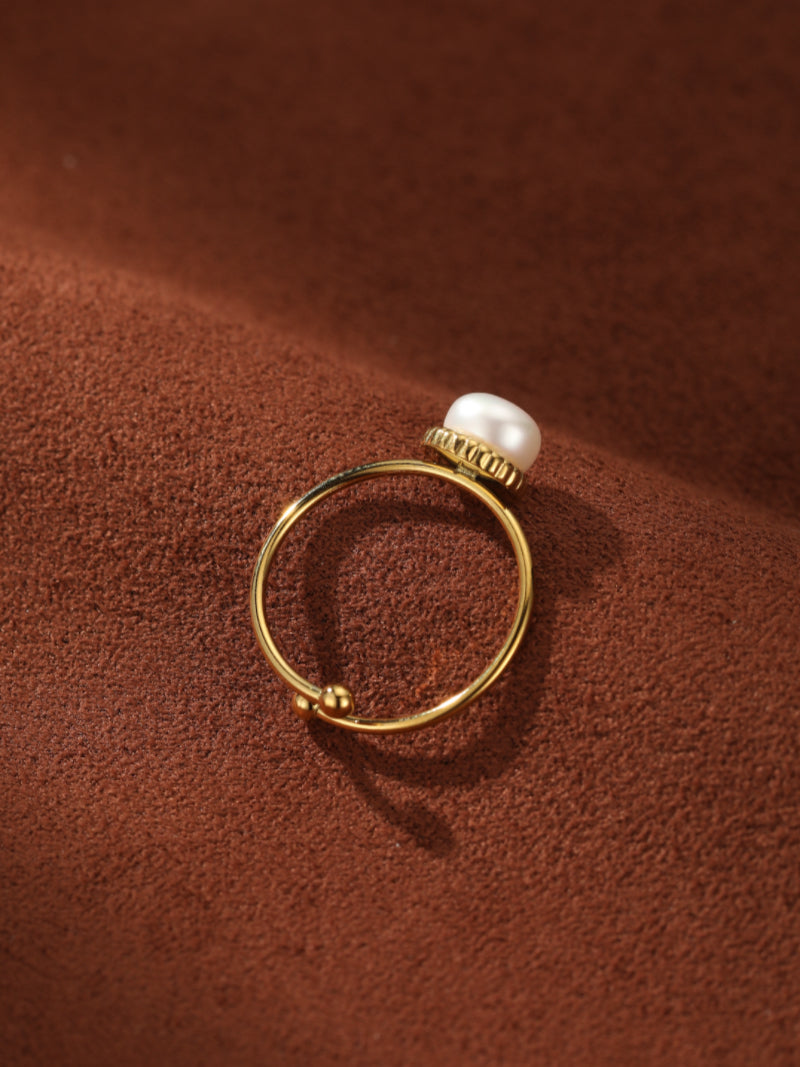 A gold-toned cuff ring with an open-ended band and a single freshwater white button pearl at the tip, set against a textured brown background.